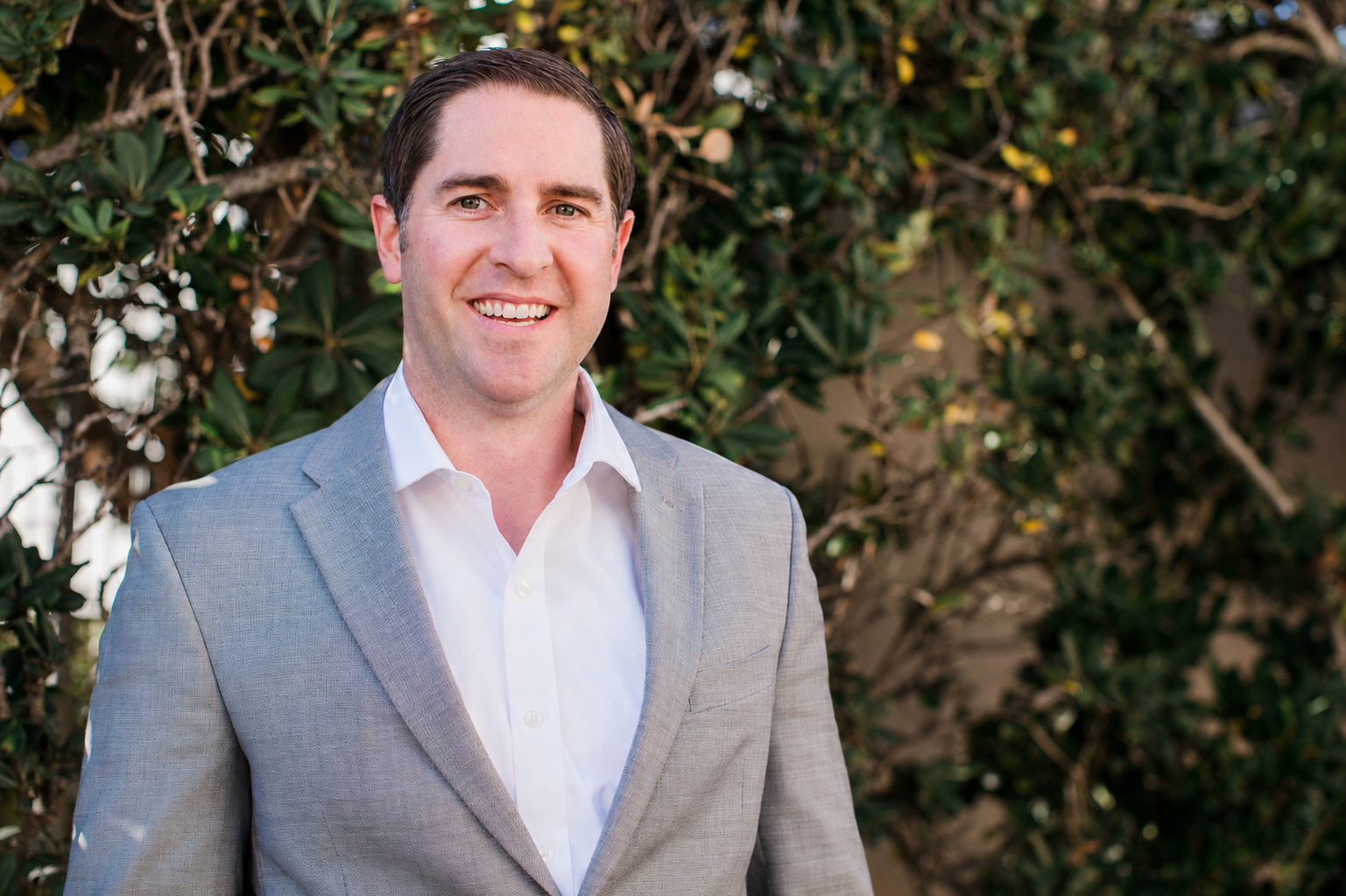A smiling man wearing a light gray suit and white shirt standing outdoors in front of green foliage.