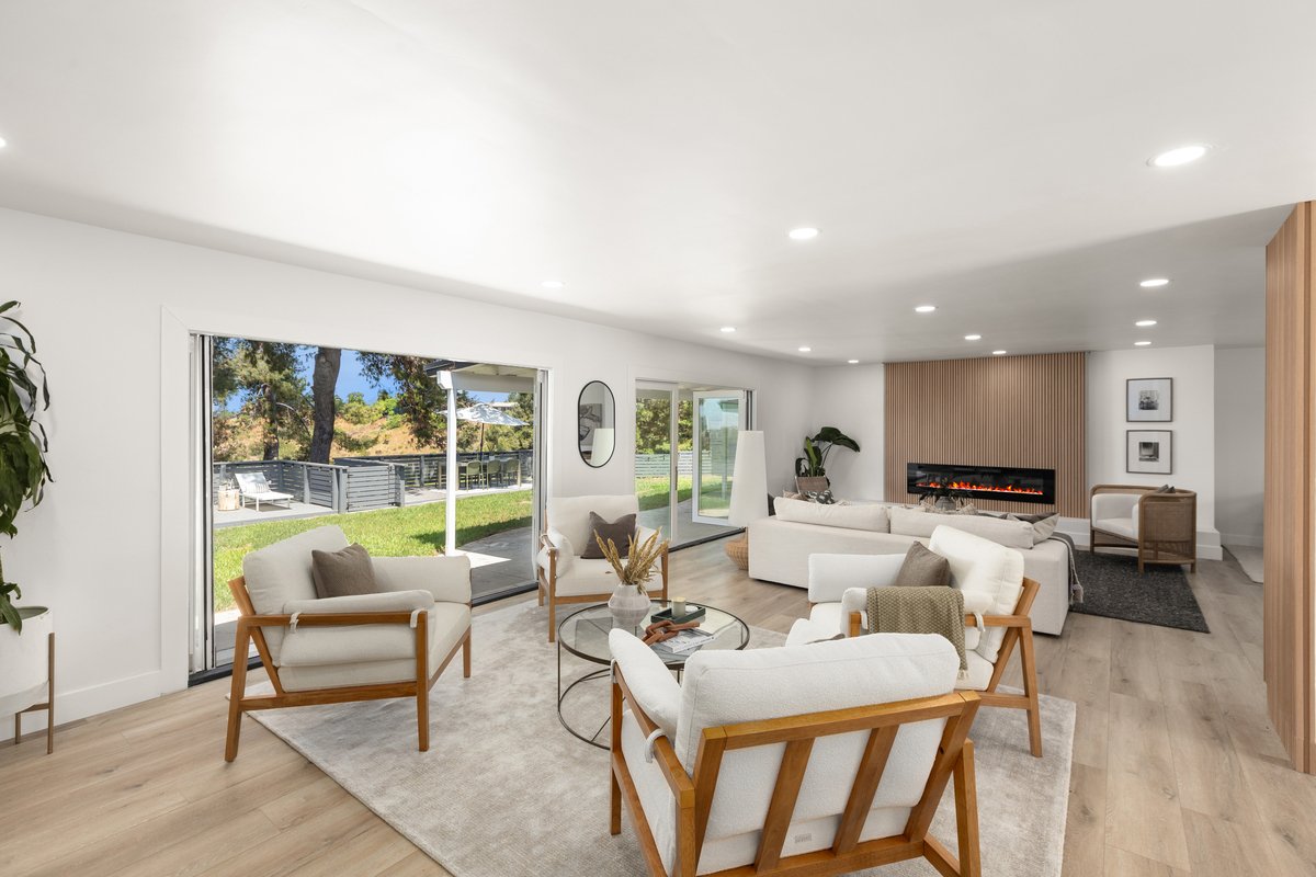 Modern living room with white sofas and armchairs with wooden frames, light hardwood flooring, a glass coffee table with decor, a fireplace, and large sliding glass doors opening to a backyard with grass and trees.