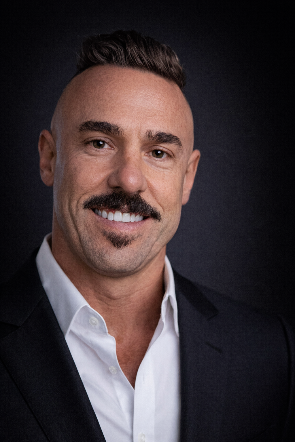 Close-up portrait of a man smiling, with a trimmed beard, mustache, and trendy hairstyle, wearing a white shirt and black blazer against a dark background.