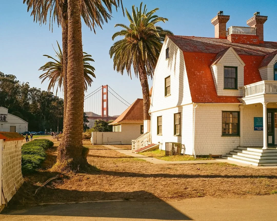 A white house with a red-tiled roof and a front porch, situated next to tall palm trees. The Golden Gate Bridge is visible in the background on a clear day.
