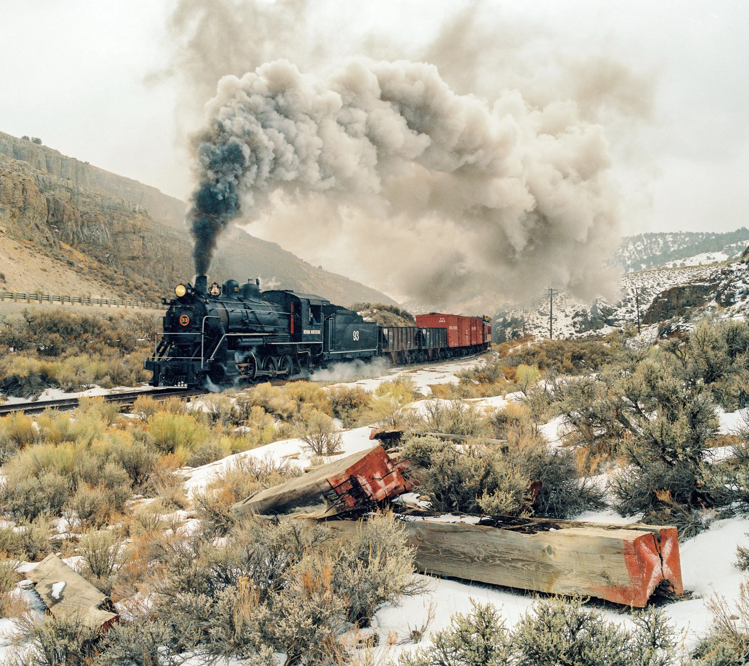 A black steam locomotive pulling red freight cars through a snowy, mountainous desert landscape with sparse vegetation and overturned train cars in the foreground, and large puffs of smoke in the sky.