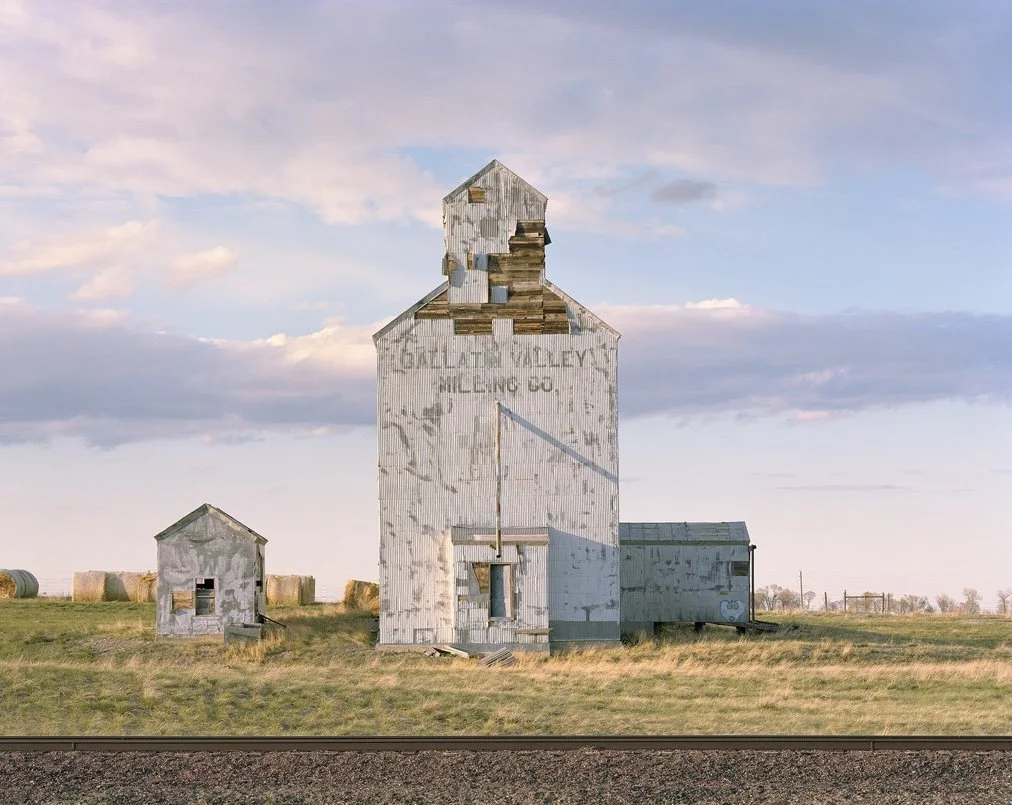 An old, weathered grain elevator with peeling paint and few windows in a rural field, surrounded by hay bales under an cloudy sky.