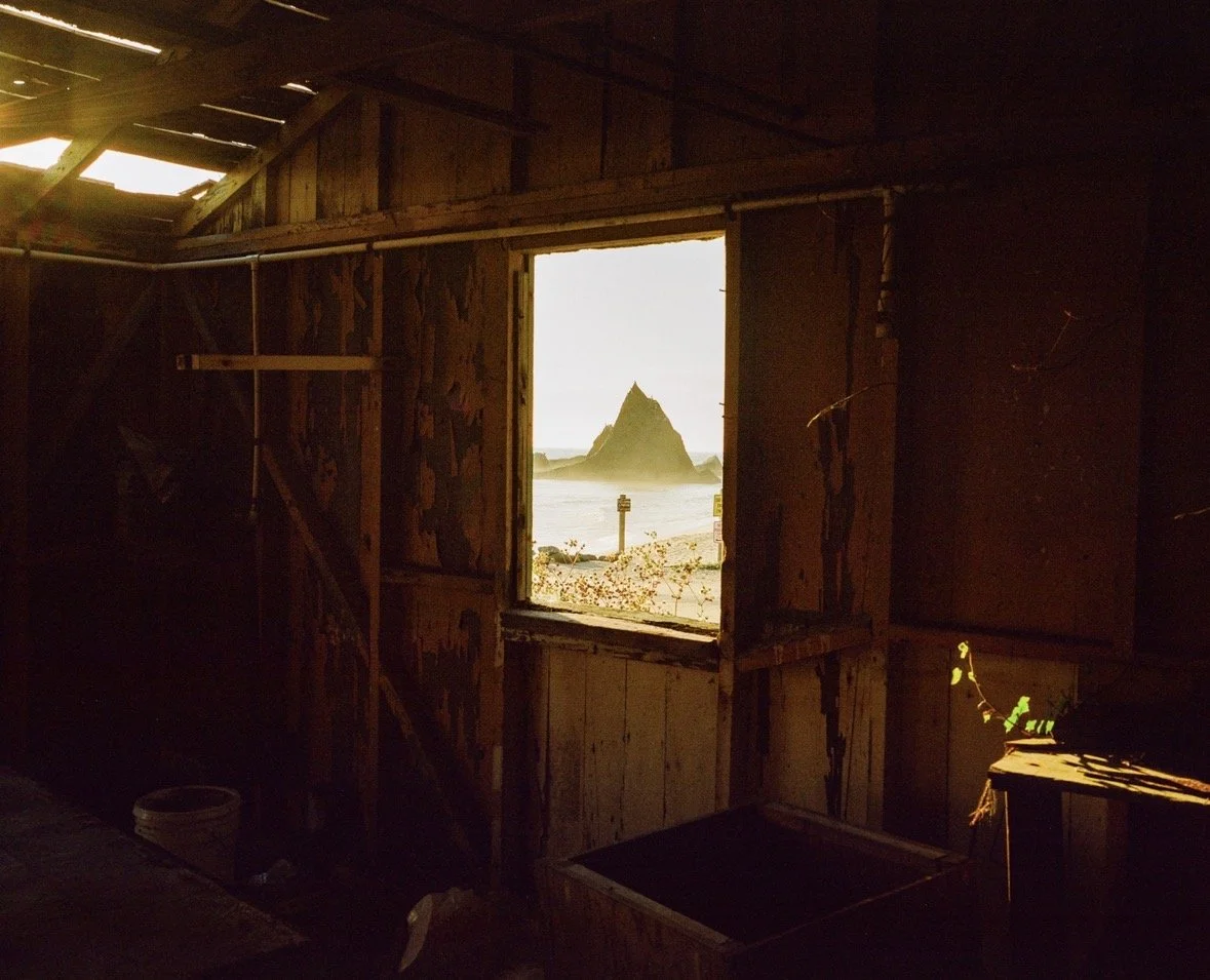 View of a rocky island with a pointed peak seen through a window in a wooden, dilapidated interior building, possibly a cabin near the beach.
