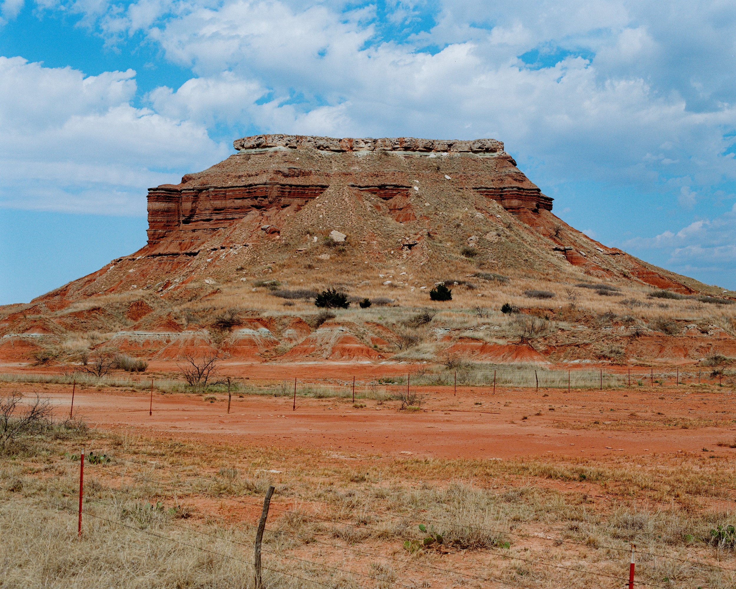 A red sandstone mesa in a desert landscape with sparse vegetation under a partly cloudy sky.