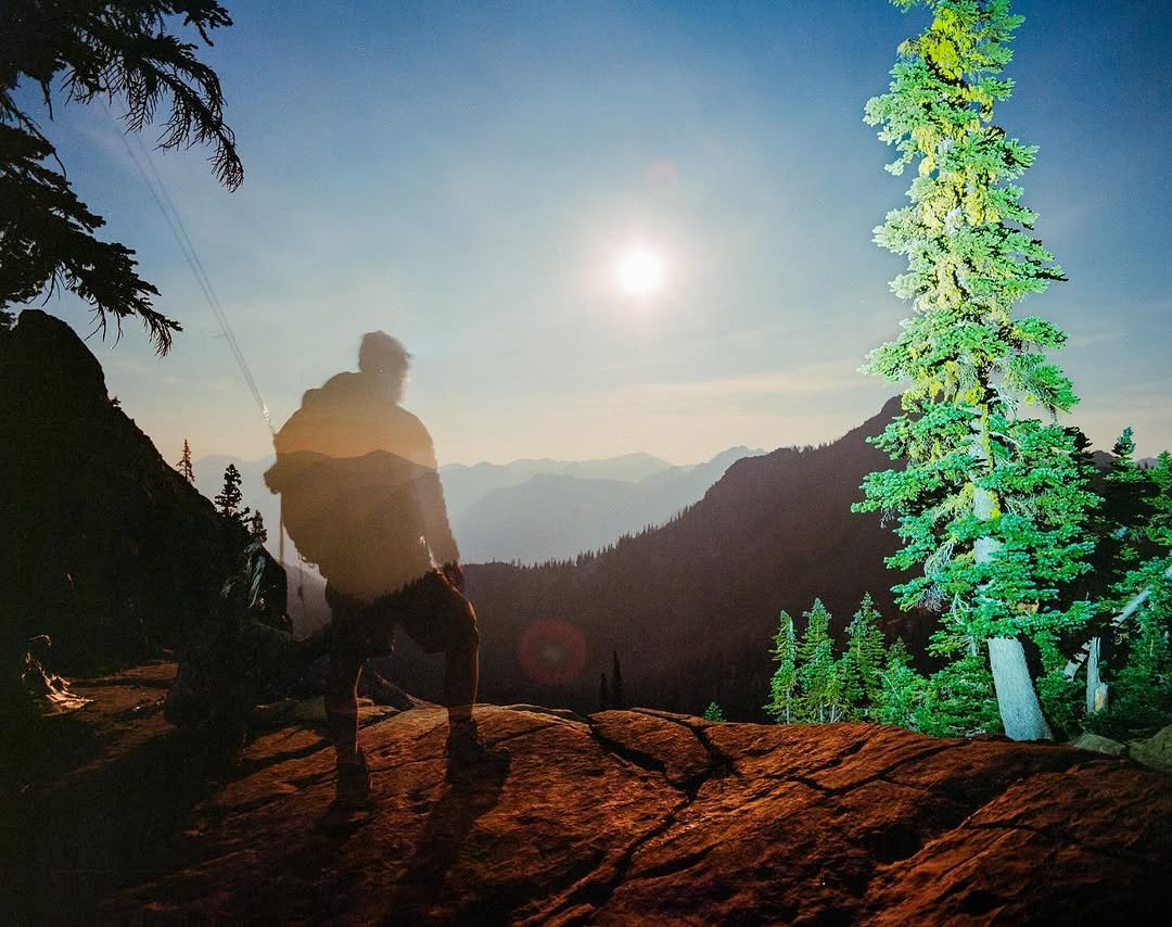 A silhouette of a person sitting on a rock at sunset in a forested mountain landscape with a full moon and illuminated green trees.