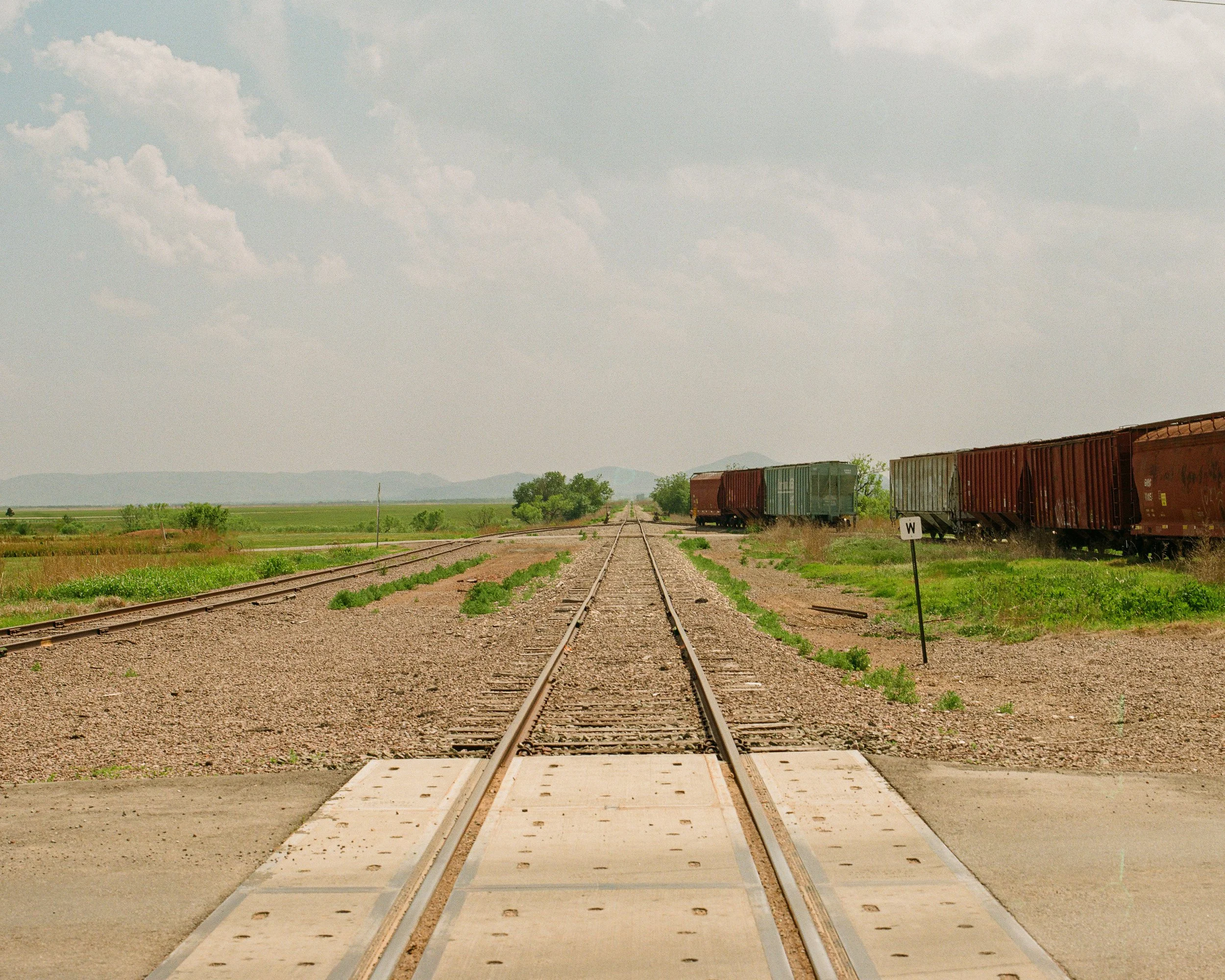 Train tracks extending into the distance with a small crossing at the front, surrounded by green fields and some railroad cars on the right side under a cloudy sky.