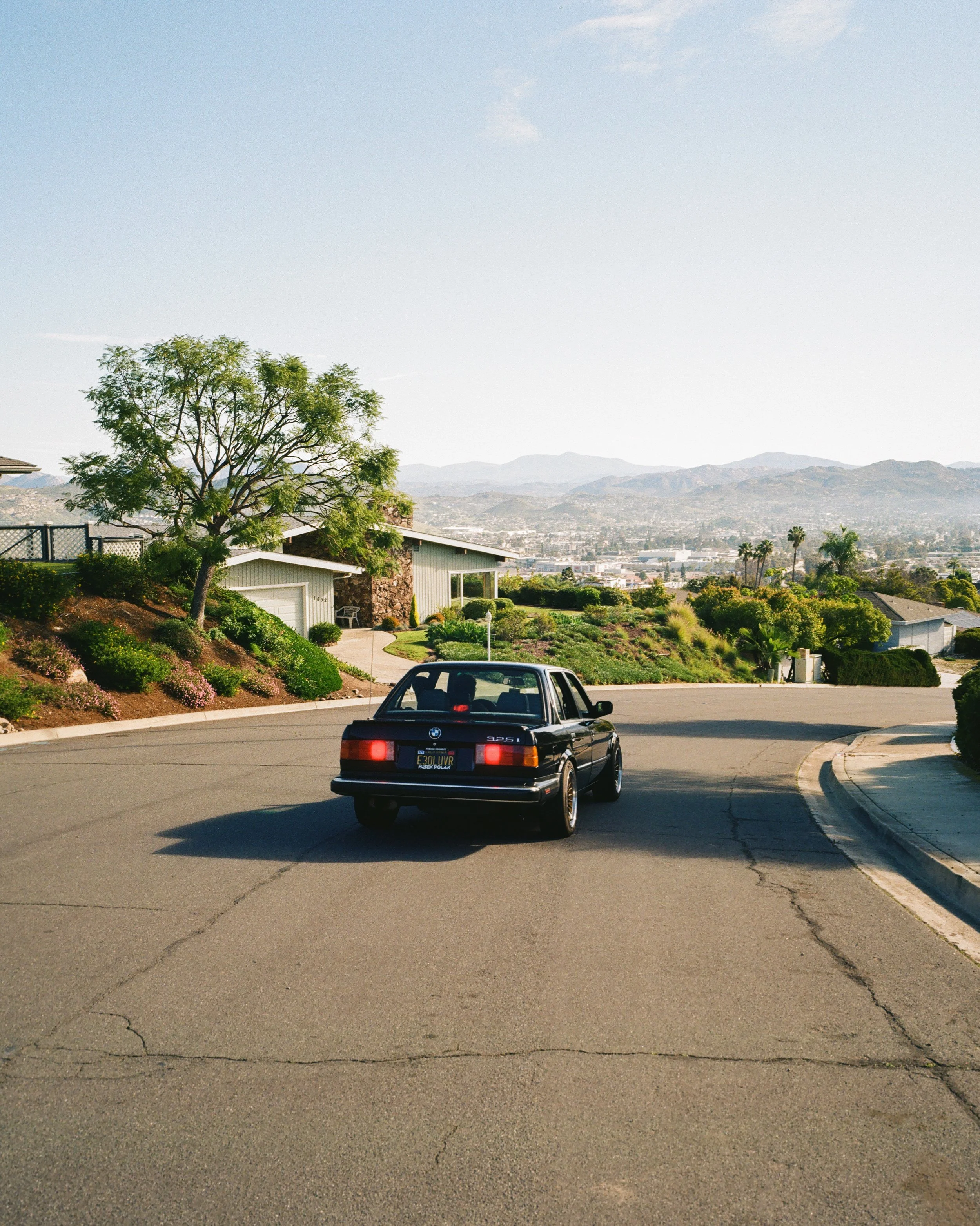 A black BMW car driving down a suburban hill on a sunny day, with houses, trees, and mountains in the background.