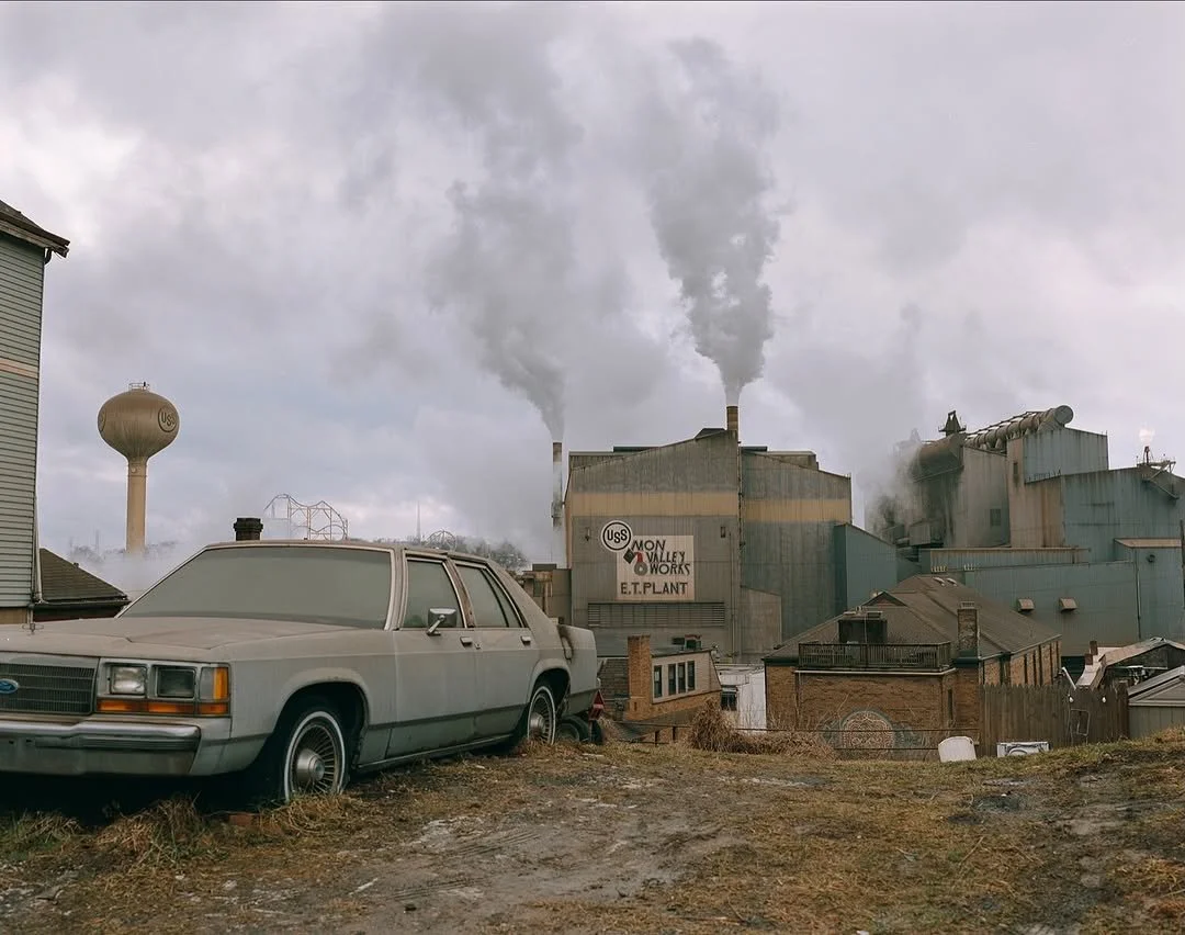 An industrial factory with smoke billowing from chimneys, a water tower nearby, and an old parked car in the foreground.