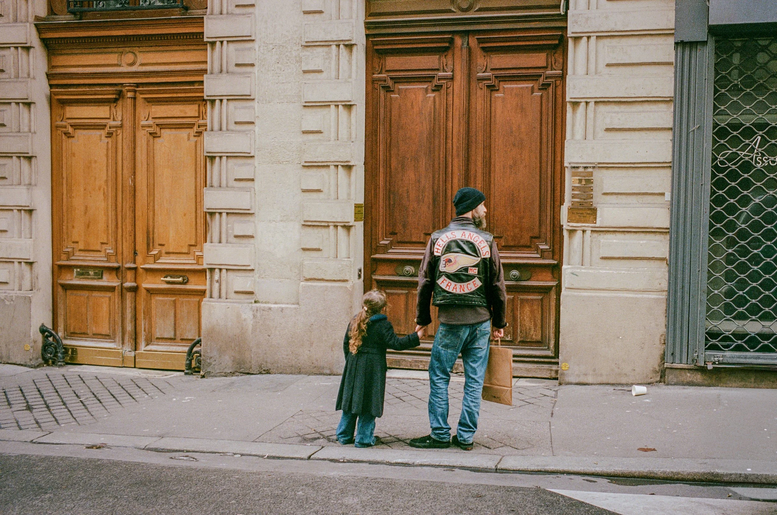 A man and a young girl holding hands on the sidewalk, waiting in front of wooden doors. The man is wearing a black beanie, a leather vest with an 'Hells Angels France' patch, and blue jeans. The girl is wearing a dark coat and blue pants.