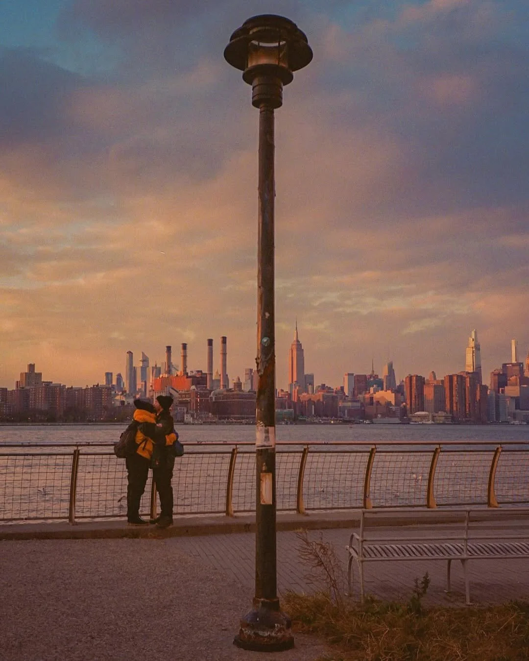Two people stand close together on a riverside promenade with a city skyline in the background, during sunset, under an old lamp post.