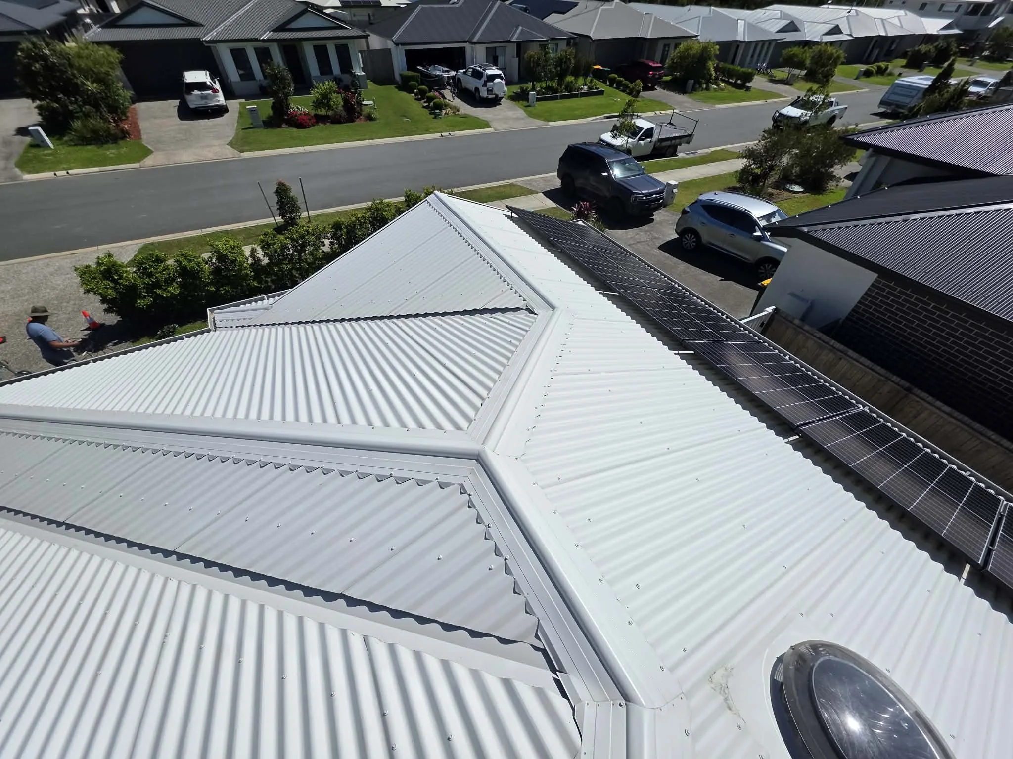 Aerial view of white metal roofing with solar panels on one section of a house, overlooking a suburban street with parked cars, houses, and greenery.