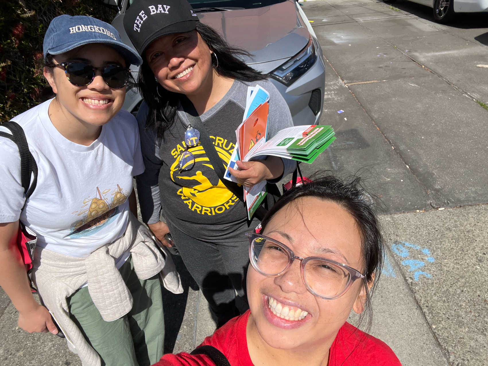 Three women smiling and taking a selfie outdoors on a sunny day, with cars and pavement in the background.