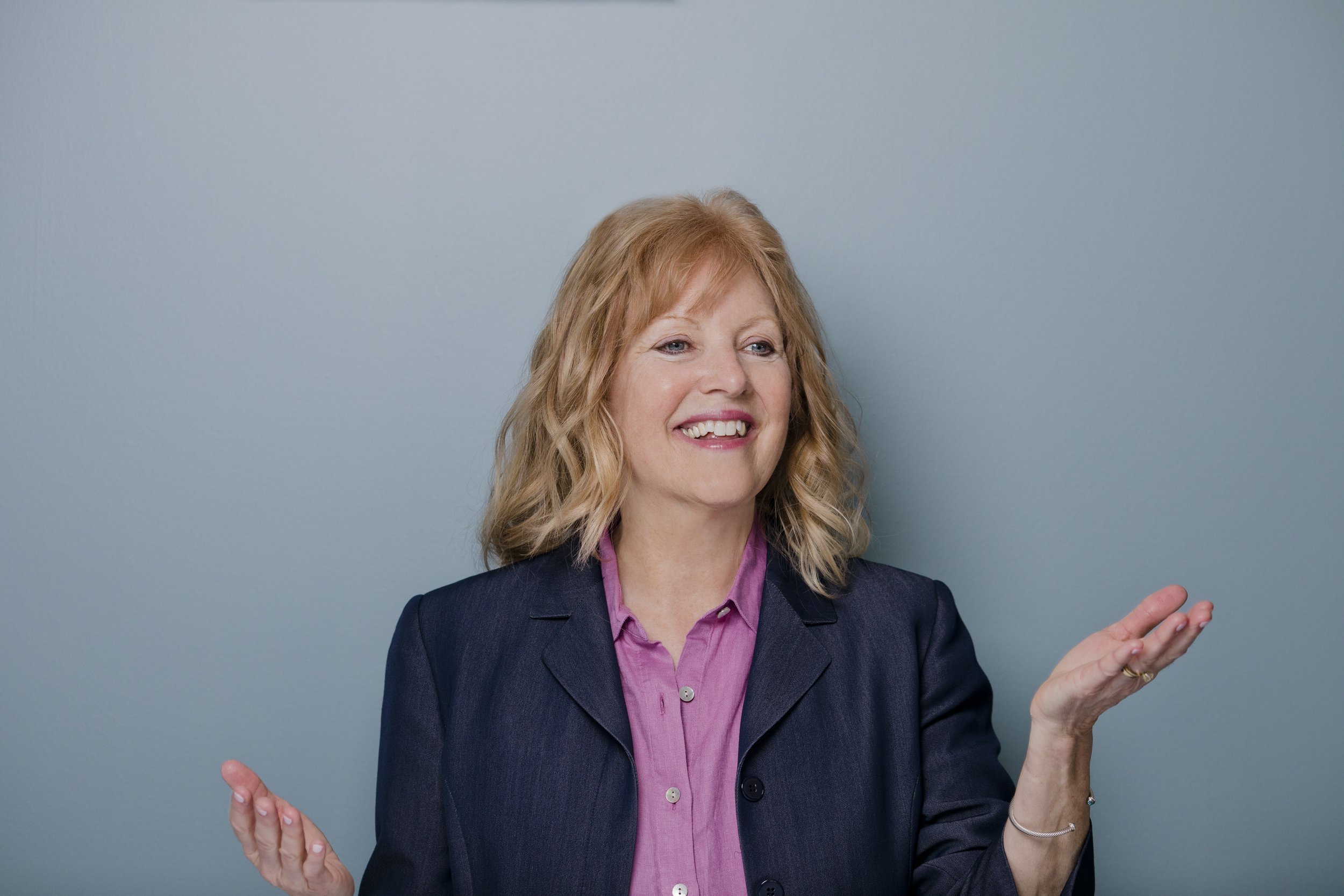 A middle-aged woman with wavy blonde hair, wearing a dark blazer over a pink shirt, is smiling and gesturing with her hands in front of a plain light blue wall.