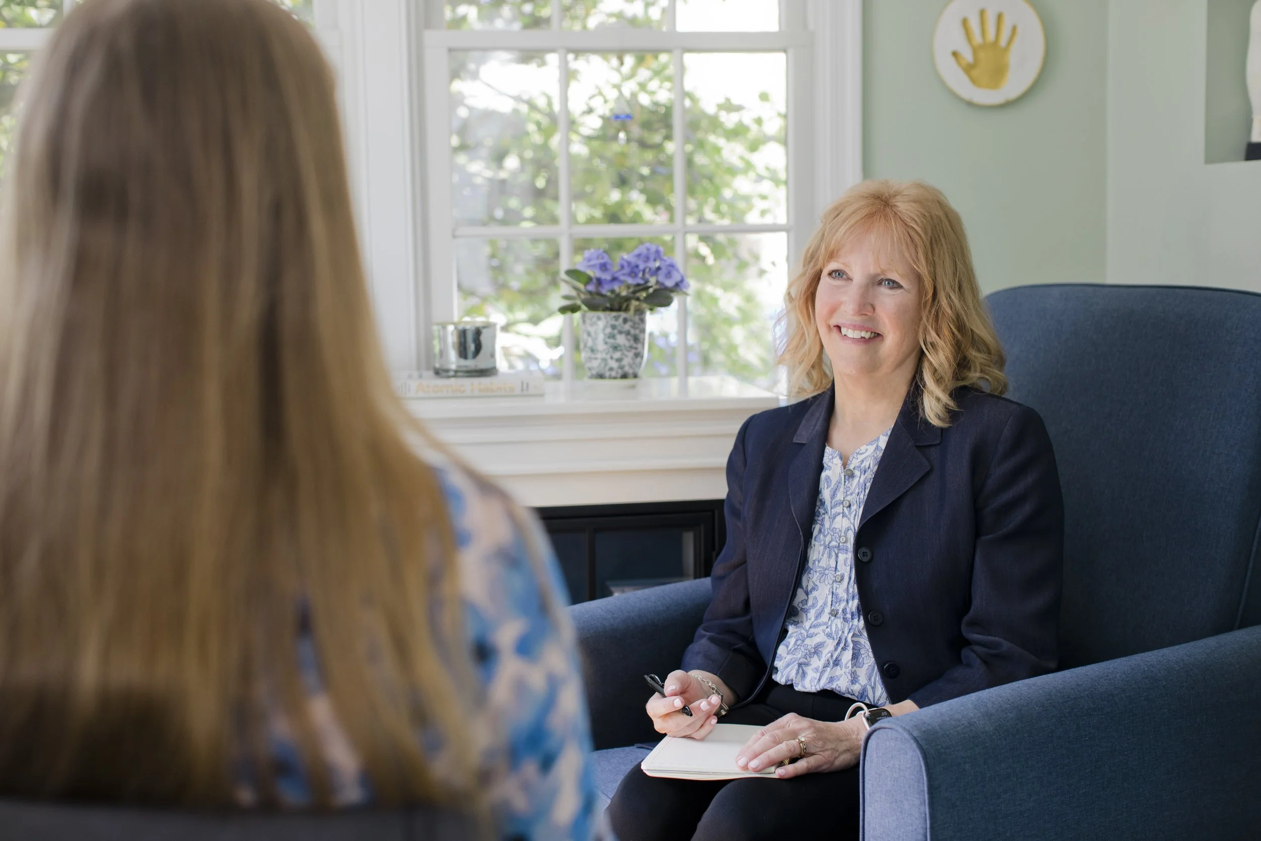 A woman with blonde hair talking to another woman whose back is facing the camera, in a room with a large window, a potted plant, and a decorative handprint on the wall.