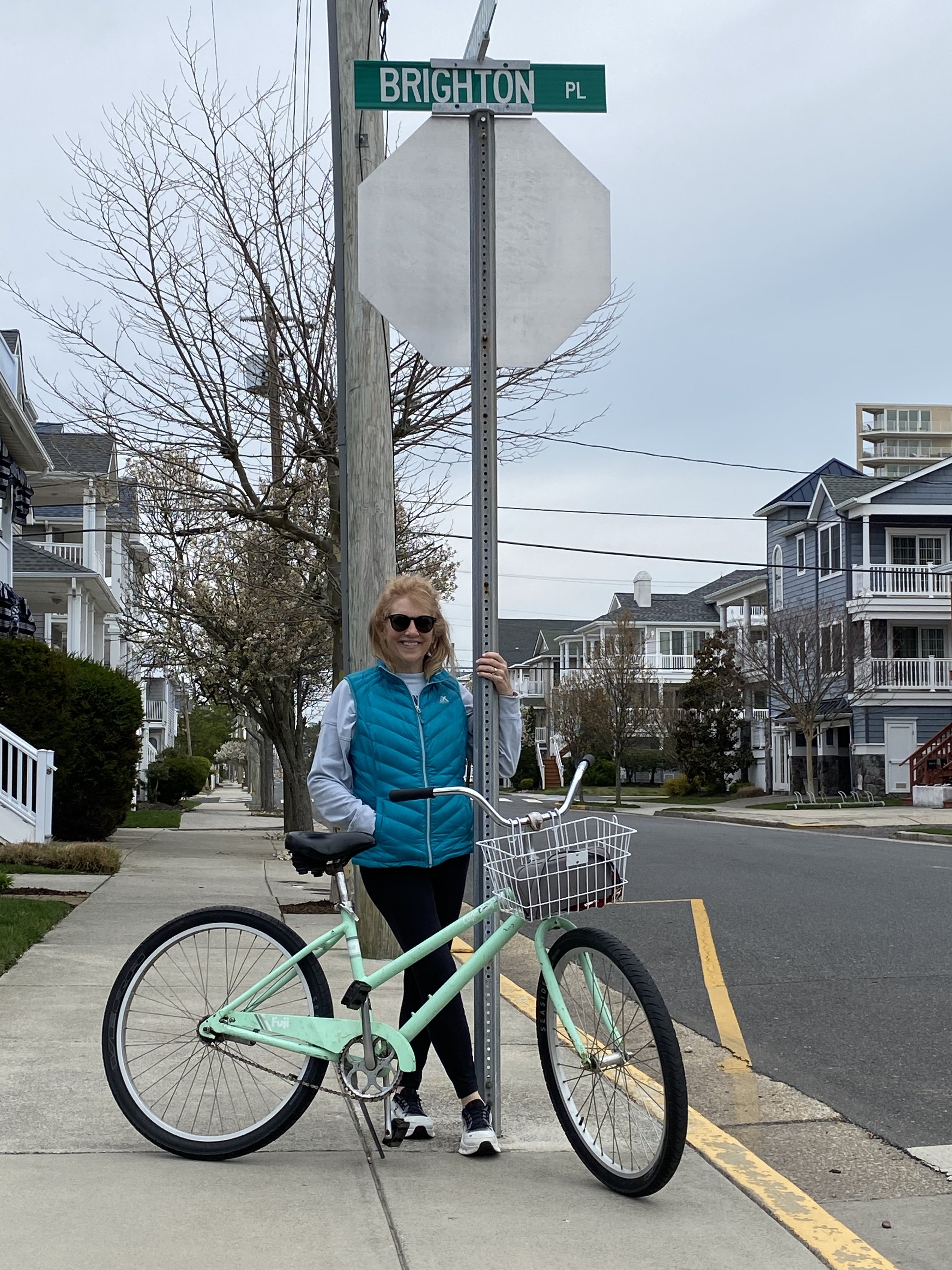 A woman with sunglasses and a bright blue vest standing next to a mint green bicycle with a basket, holding a stop sign pole at an intersection on a cloudy day.
