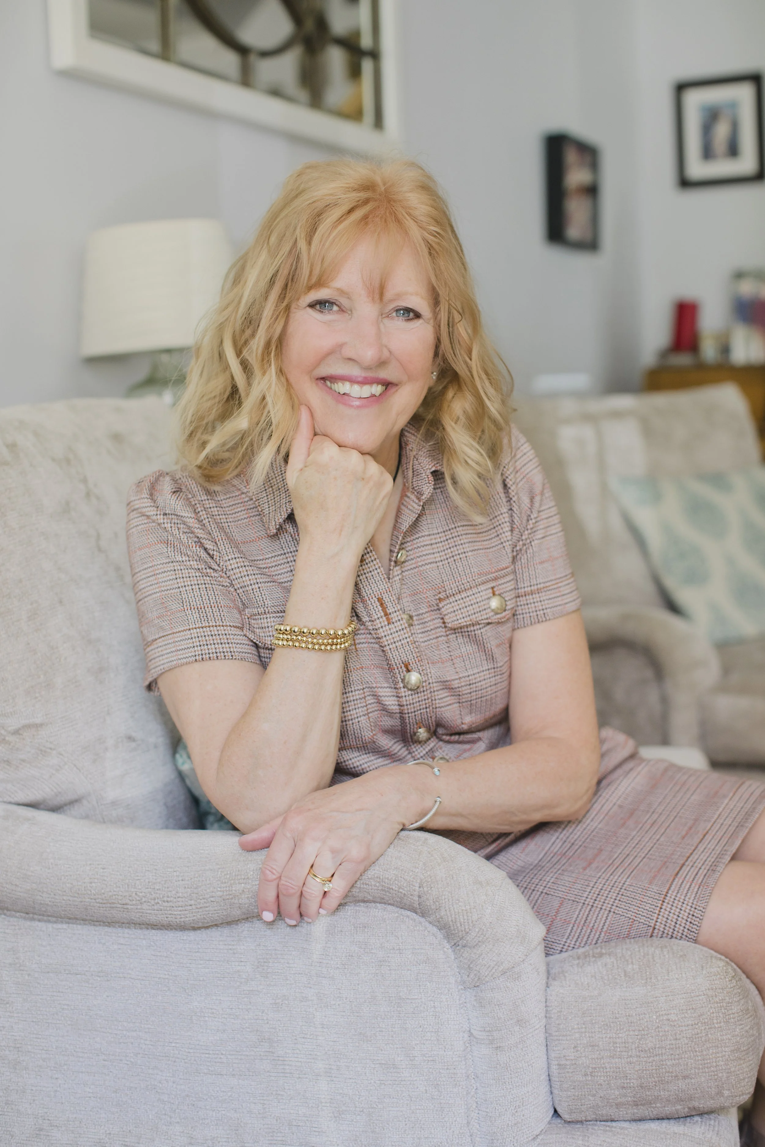A smiling woman with blonde hair and blue eyes, sitting on a beige couch in a living room, wearing a plaid dress with pearl and gold jewelry.