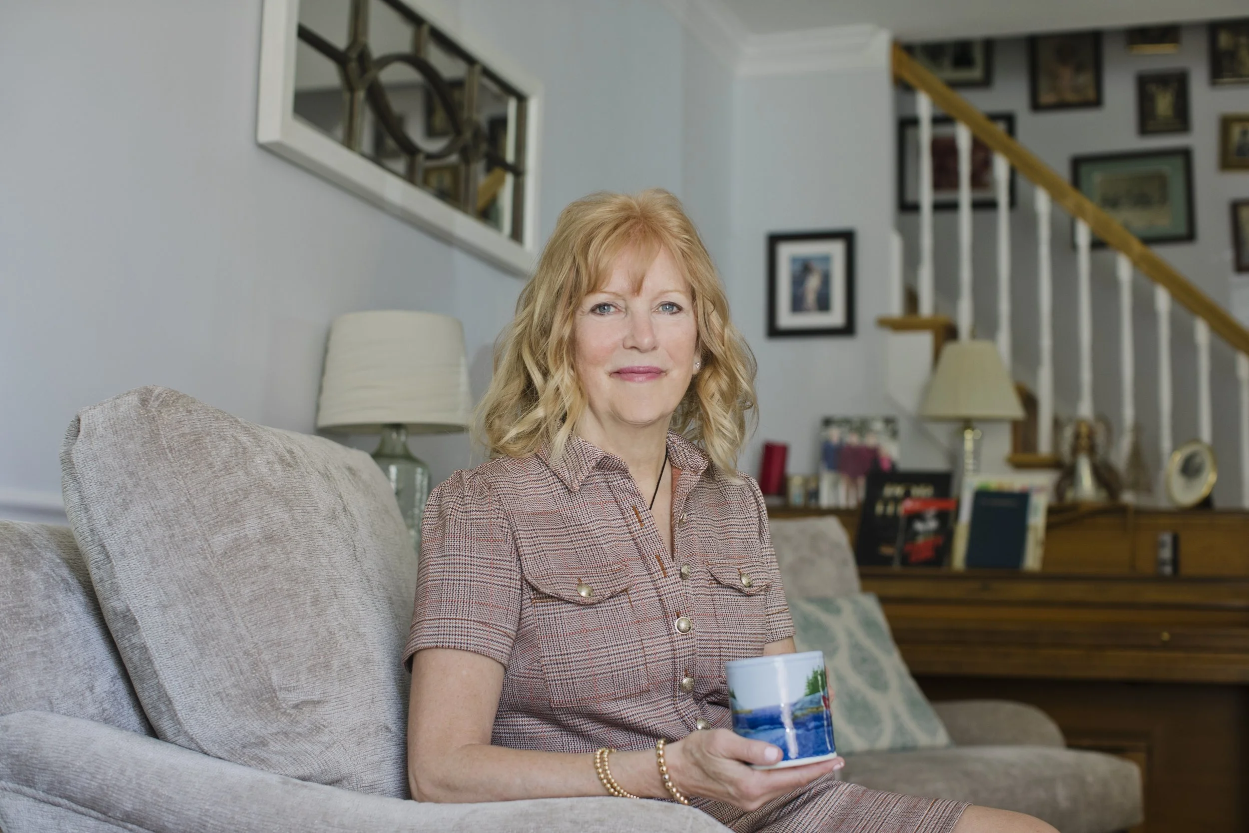 A woman with blonde hair sitting on a beige sofa, holding a colorful mug, in a living room with artwork on the wall and a staircase in the background.