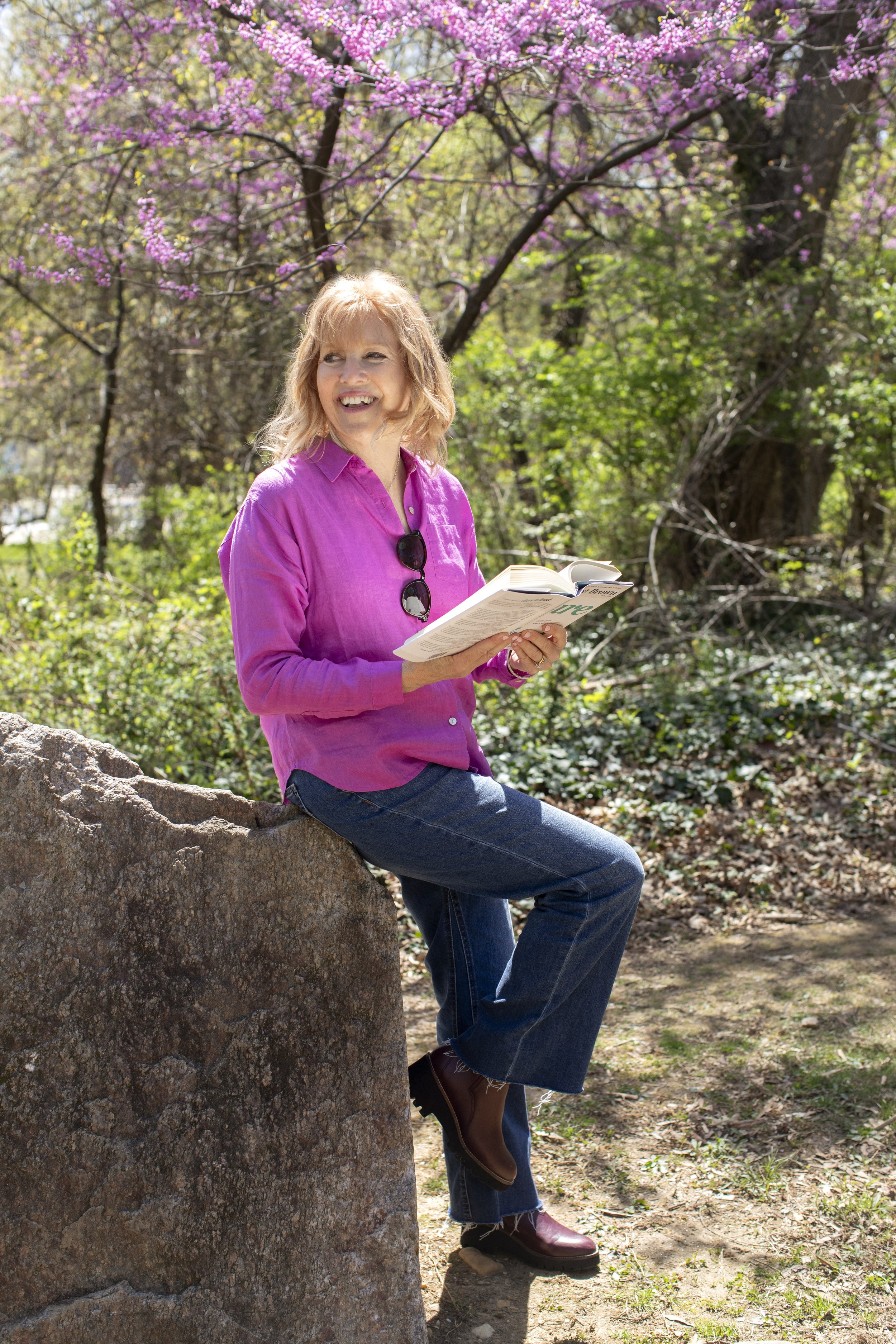 A woman with blond hair, wearing a pink shirt and jeans, sitting on a large rock outdoors among blooming trees with pink flowers, holding an open book.