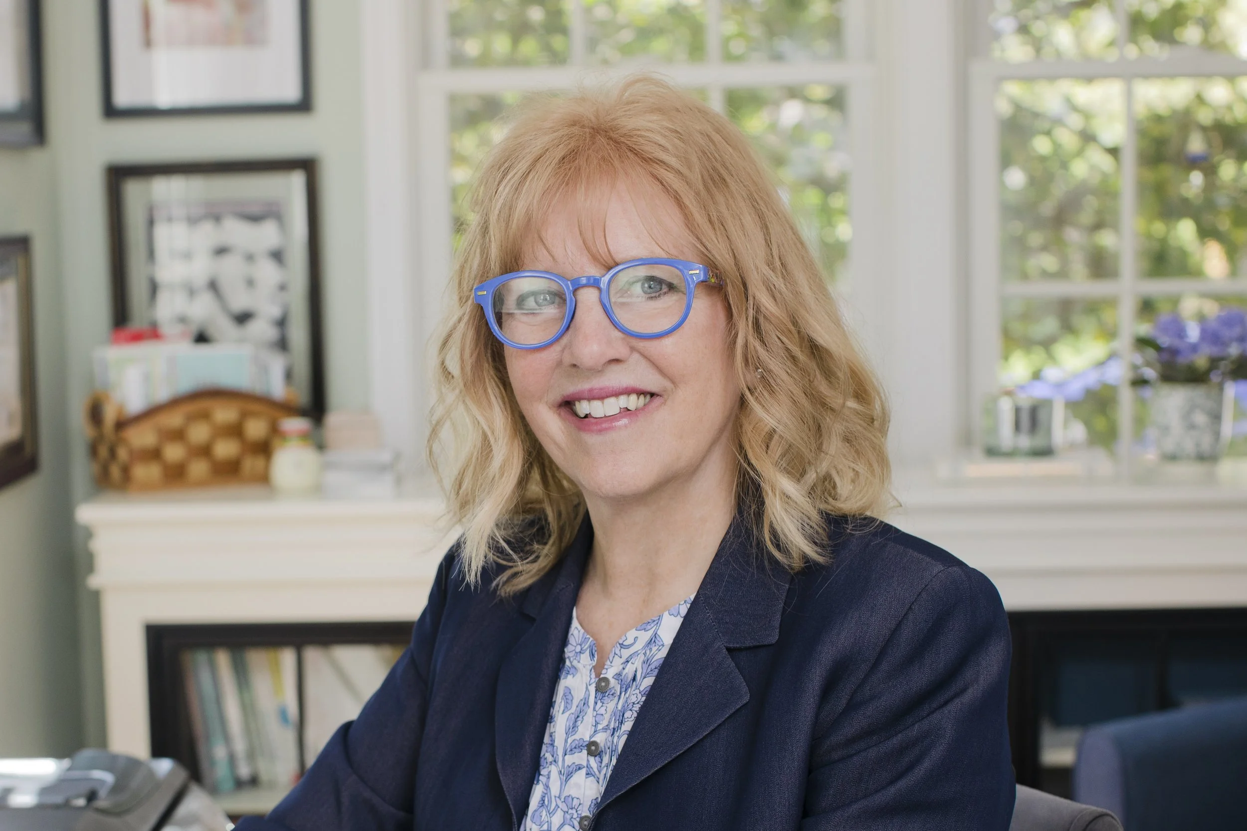 A woman with red hair, wearing blue glasses and a navy blazer, smiling while sitting at a desk in an office with large windows and a bookshelf in the background.