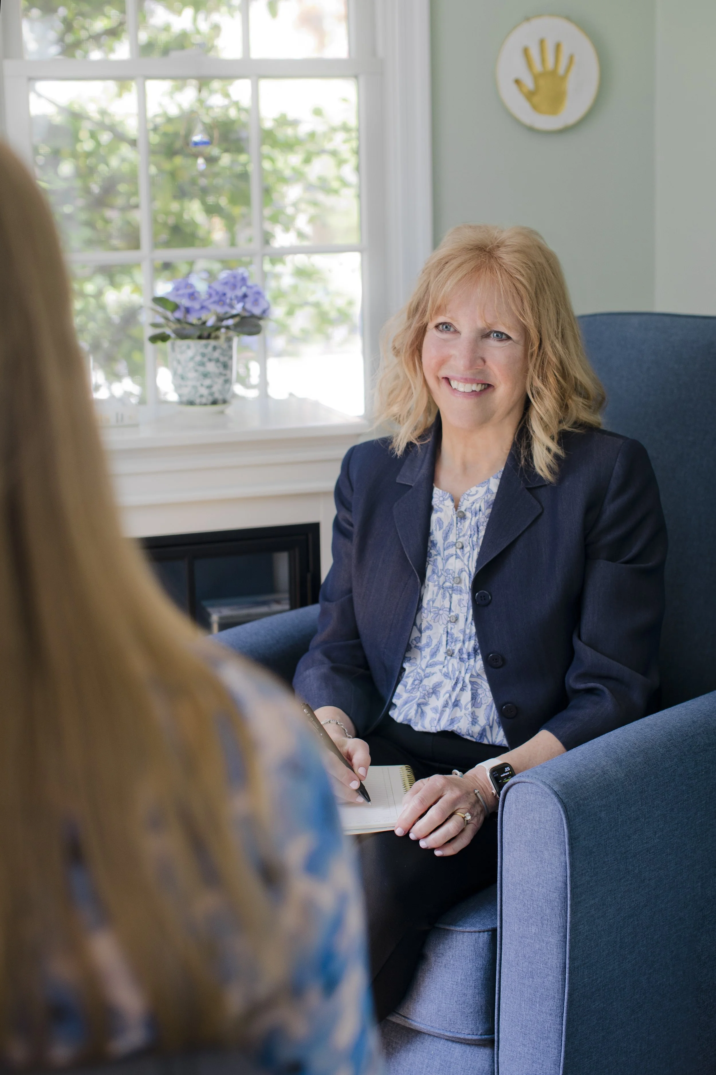 A woman with blonde hair wearing a dark blazer and a patterned blouse sitting in a blue armchair, smiling during a conversation with another person in an indoor setting with a window, a potted plant with purple flowers, and a decorative handprint on the wall in the background.