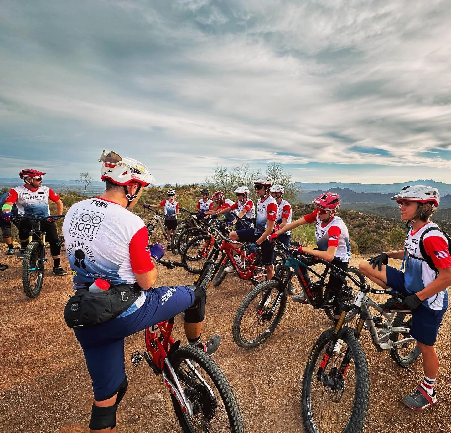 Group of cyclists in mountain biking gear and helmets gathered outdoors on a dirt trail in a desert landscape, with mountains and cloudy sky in the background.