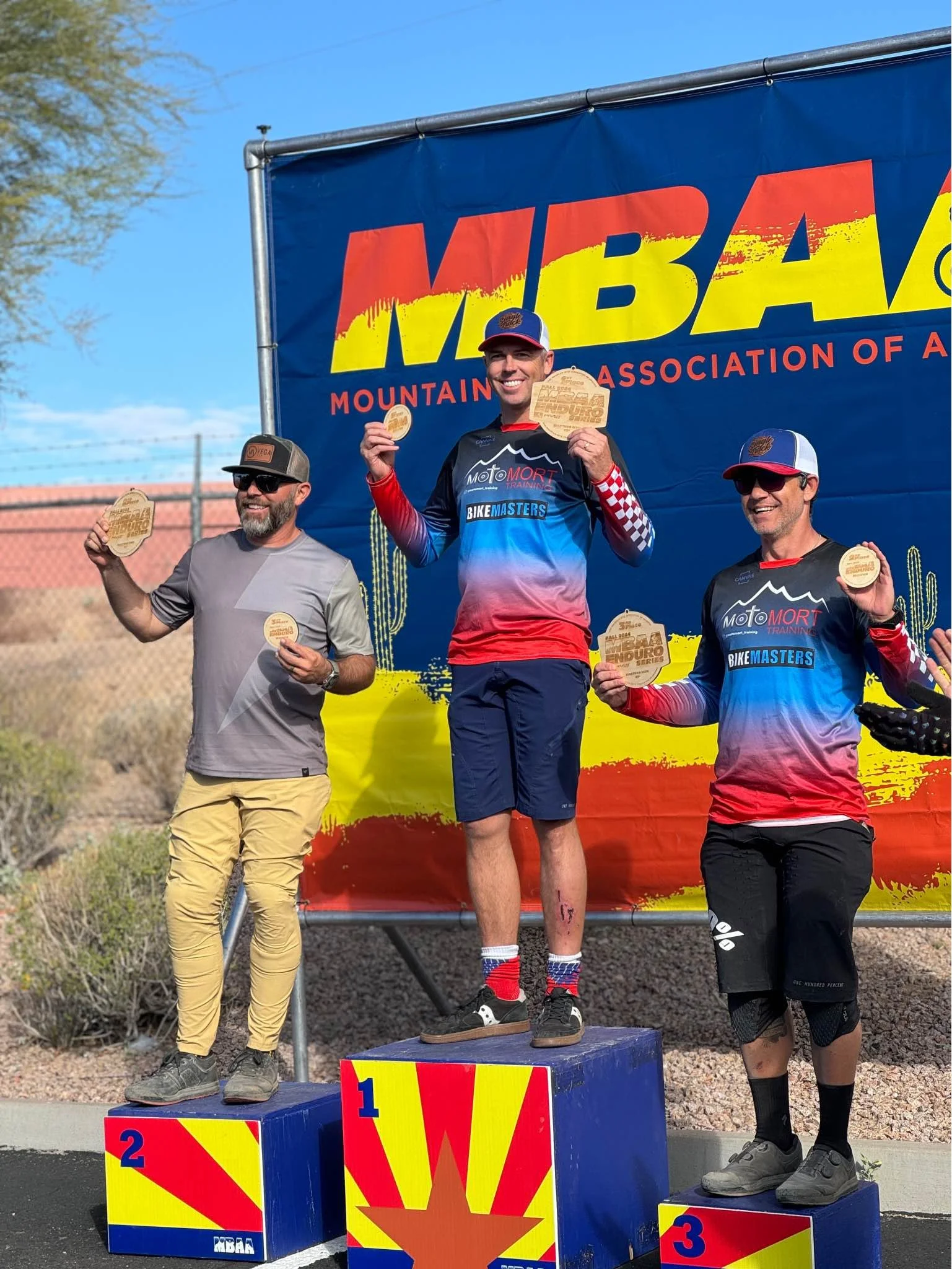 Three male mountain bikers on a winners' podium at an event hosted by the Mountain Bike Association of Arizona (MBAA), with the first-place winner in the center holding a wooden podium plaque, under a large MBAA banner with a desert landscape and blu
