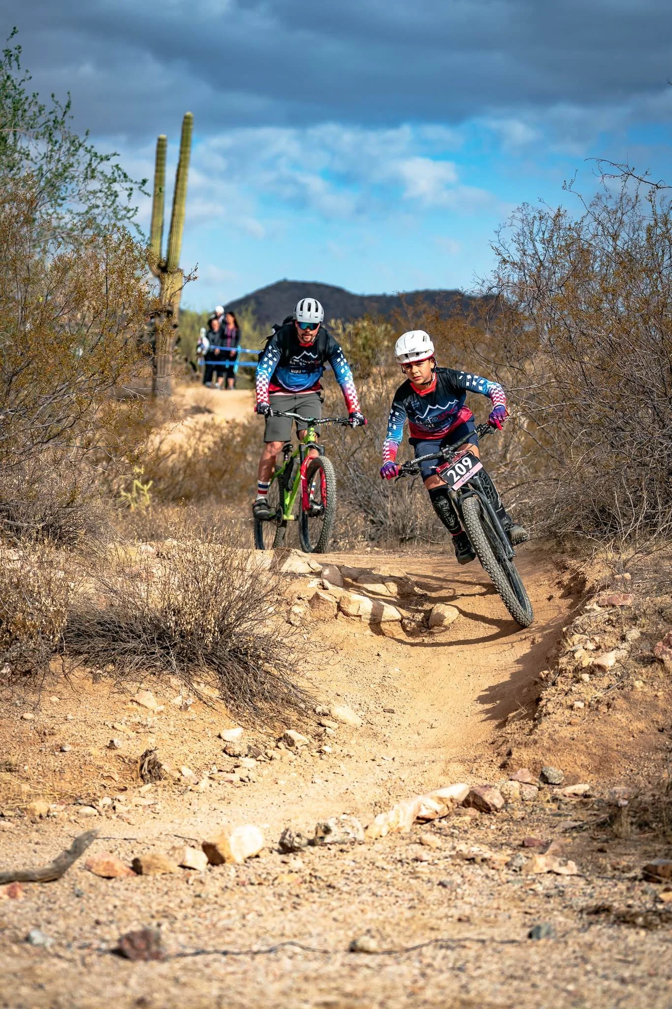 Two mountain bikers wearing helmets riding on a dirt trail through a desert landscape with cactus and bushes, under a cloudy sky.