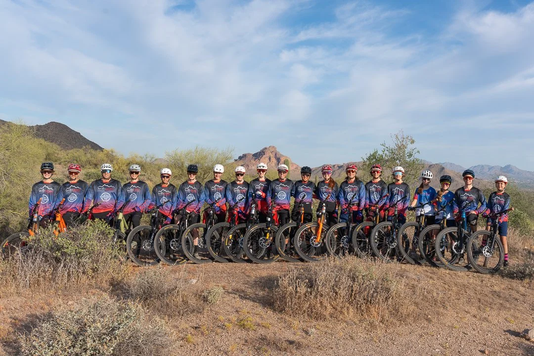 A group of cyclists in matching jerseys and helmets standing with their bikes on a desert trail with mountains in the background.