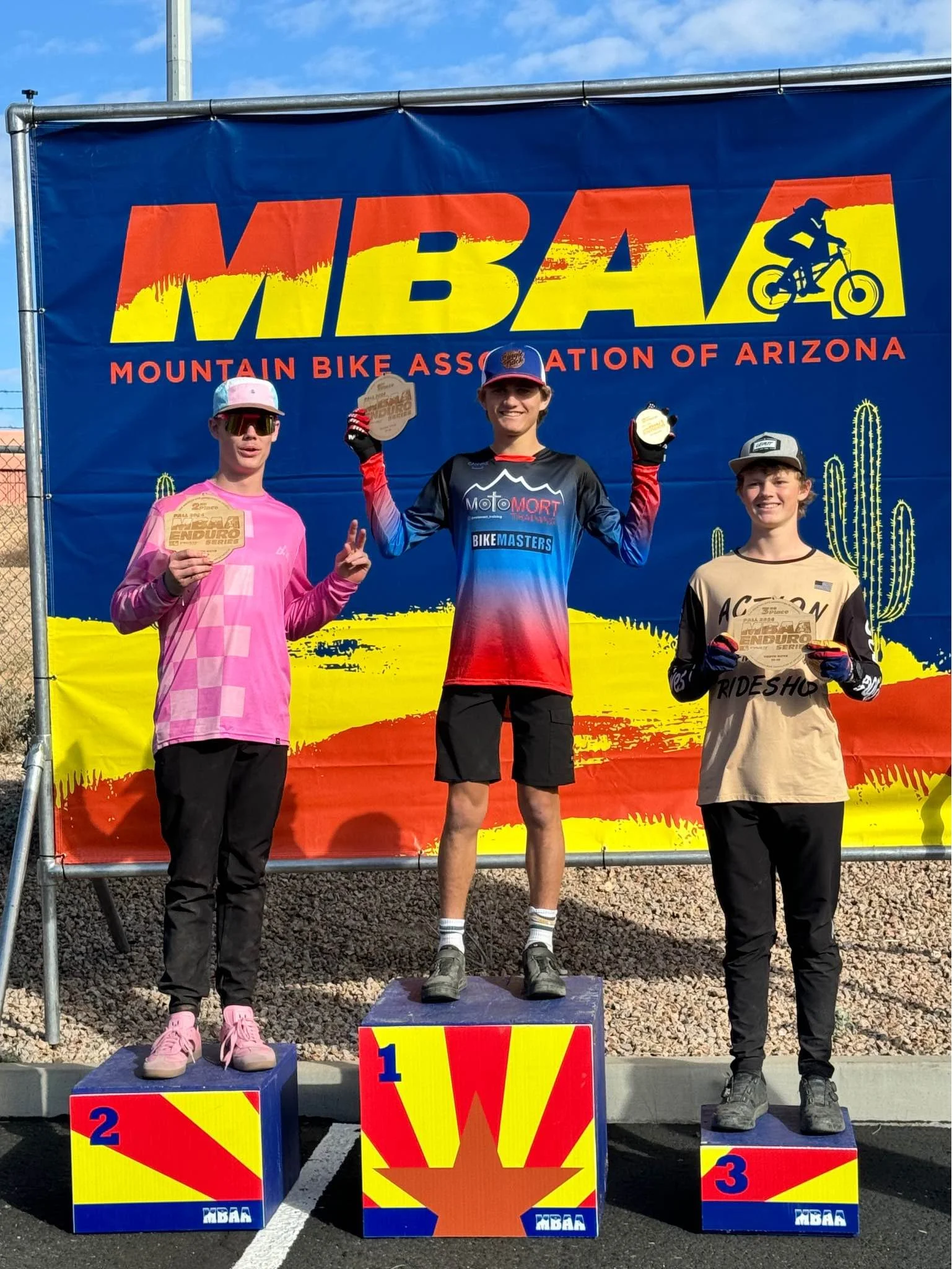 Three young male mountain bikers standing on a winners' podium, with a large banner behind them that reads "WBA Mountain Bike Association of Arizona." The boy in the center, on the first-place podium, is holding a medal and making a peace sign. The o