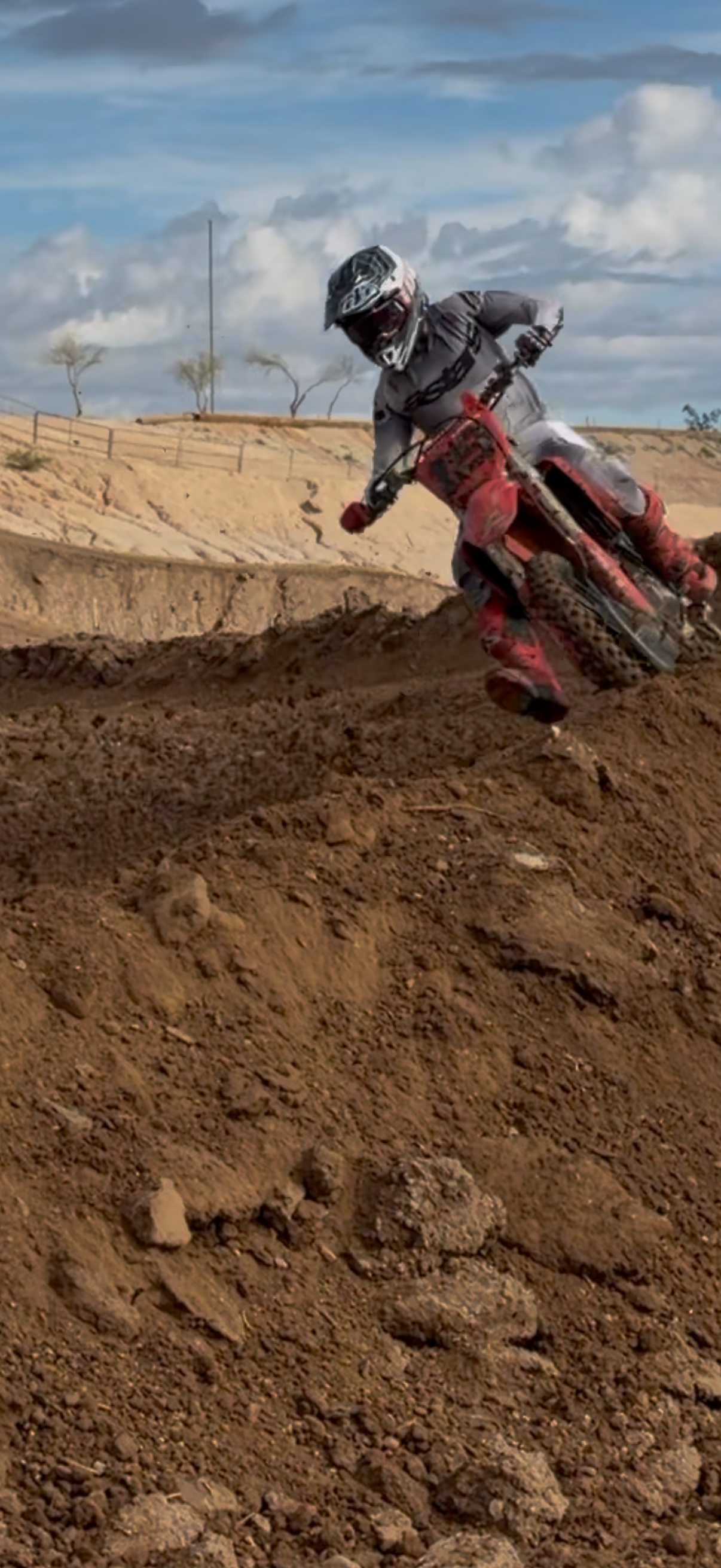 A person riding a dirt bike on a dirt track with a cloudy sky in the background.