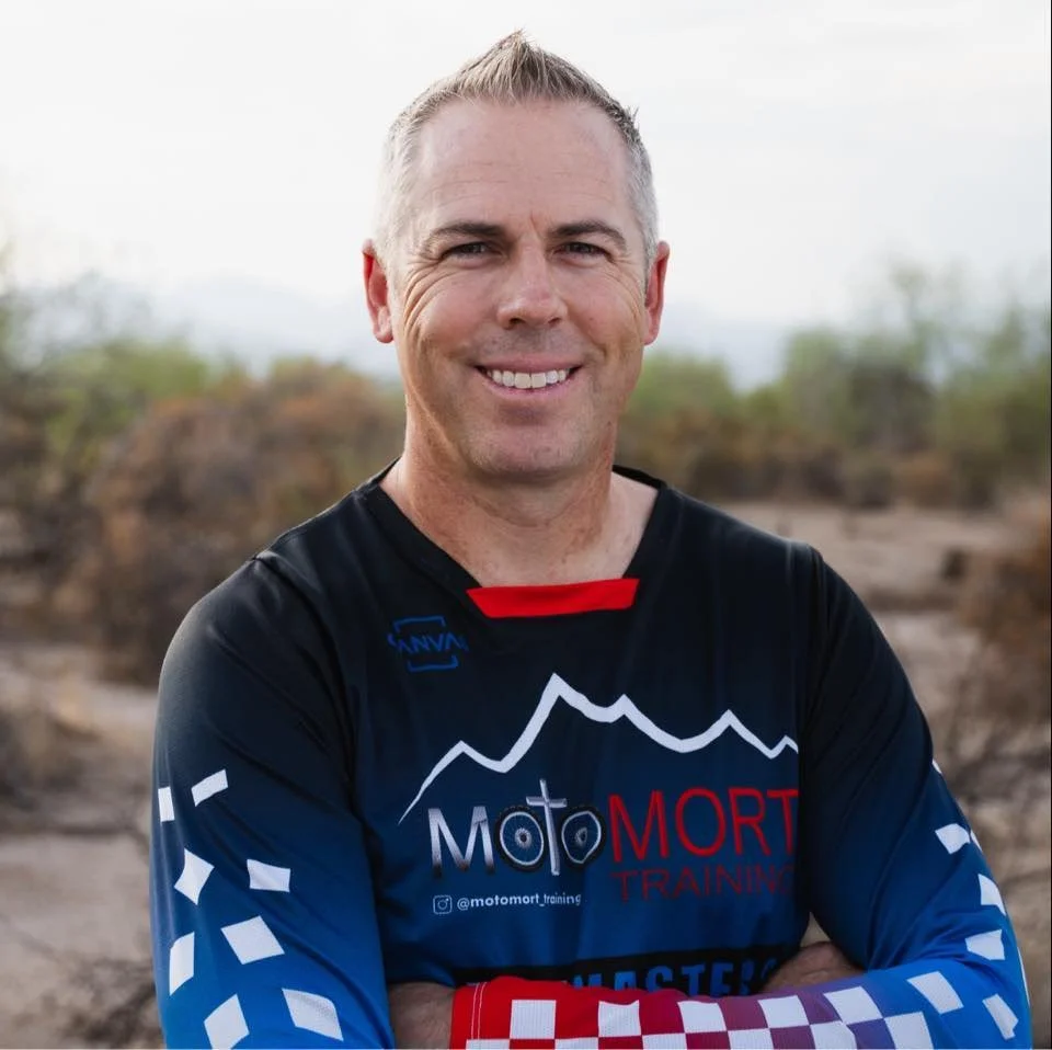 A smiling man in a black, blue, red, and white motocross jersey standing outdoors with arms crossed, with desert landscape and vegetation in the background.
