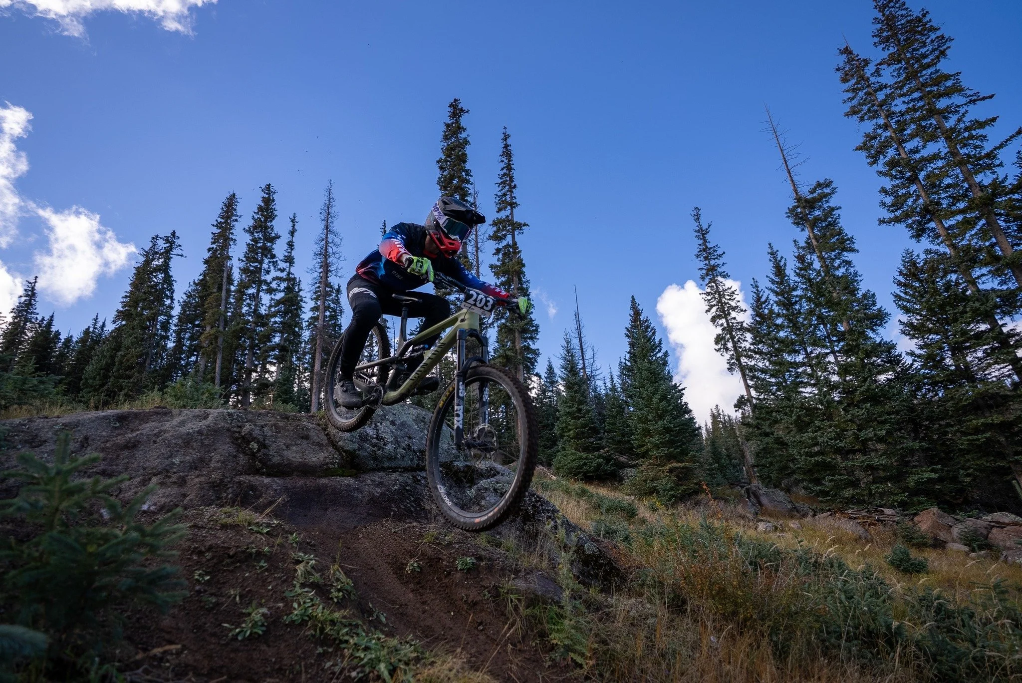 A mountain biker wearing a helmet and protective gear riding down a rocky trail in a forested area under a blue sky.