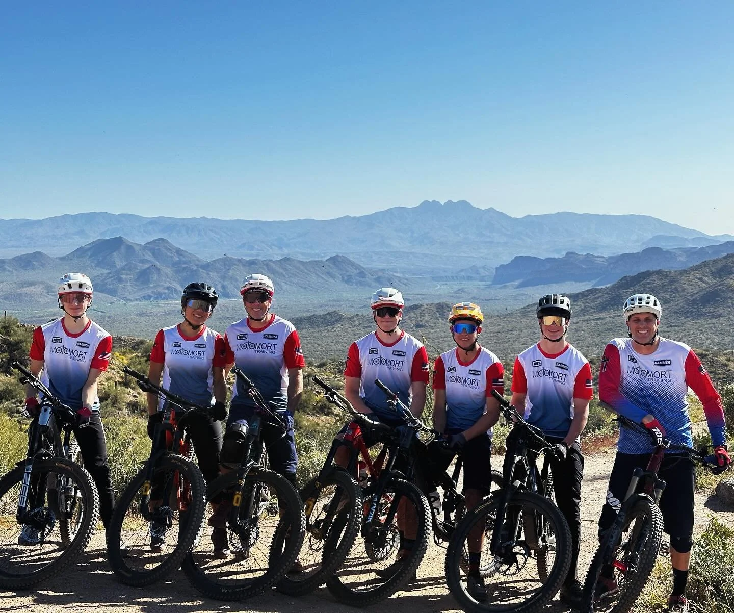 Seven cyclists in sports gear with helmets and sunglasses, standing upright with their mountain bikes in a desert landscape with mountains in the background.