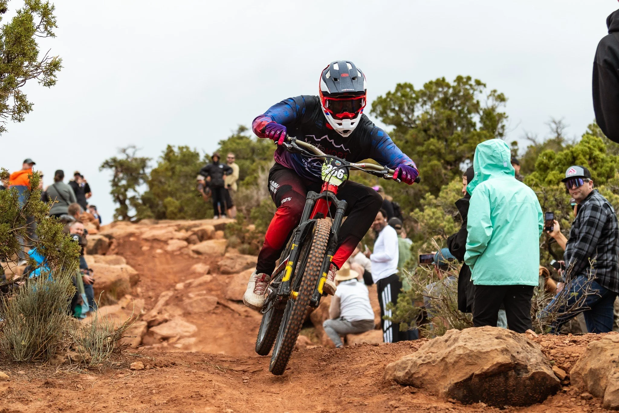 A mountain biker in a black helmet, black and blue jersey, and black and red pants jumps off a dirt trail with rocks, surrounded by spectators on a cloudy day.