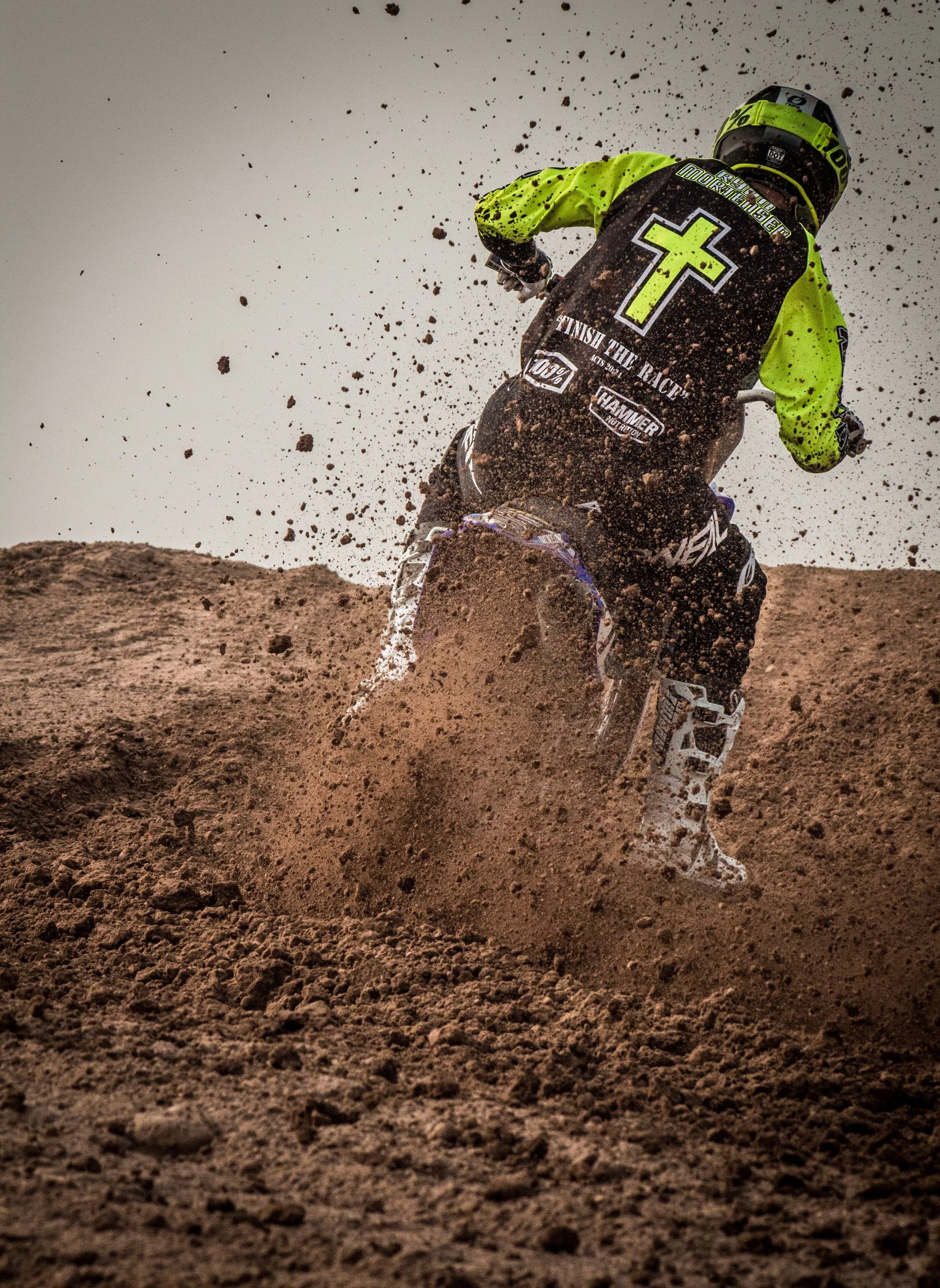 Motocross rider in black and green gear riding through dirt, causing a splash of mud behind them.