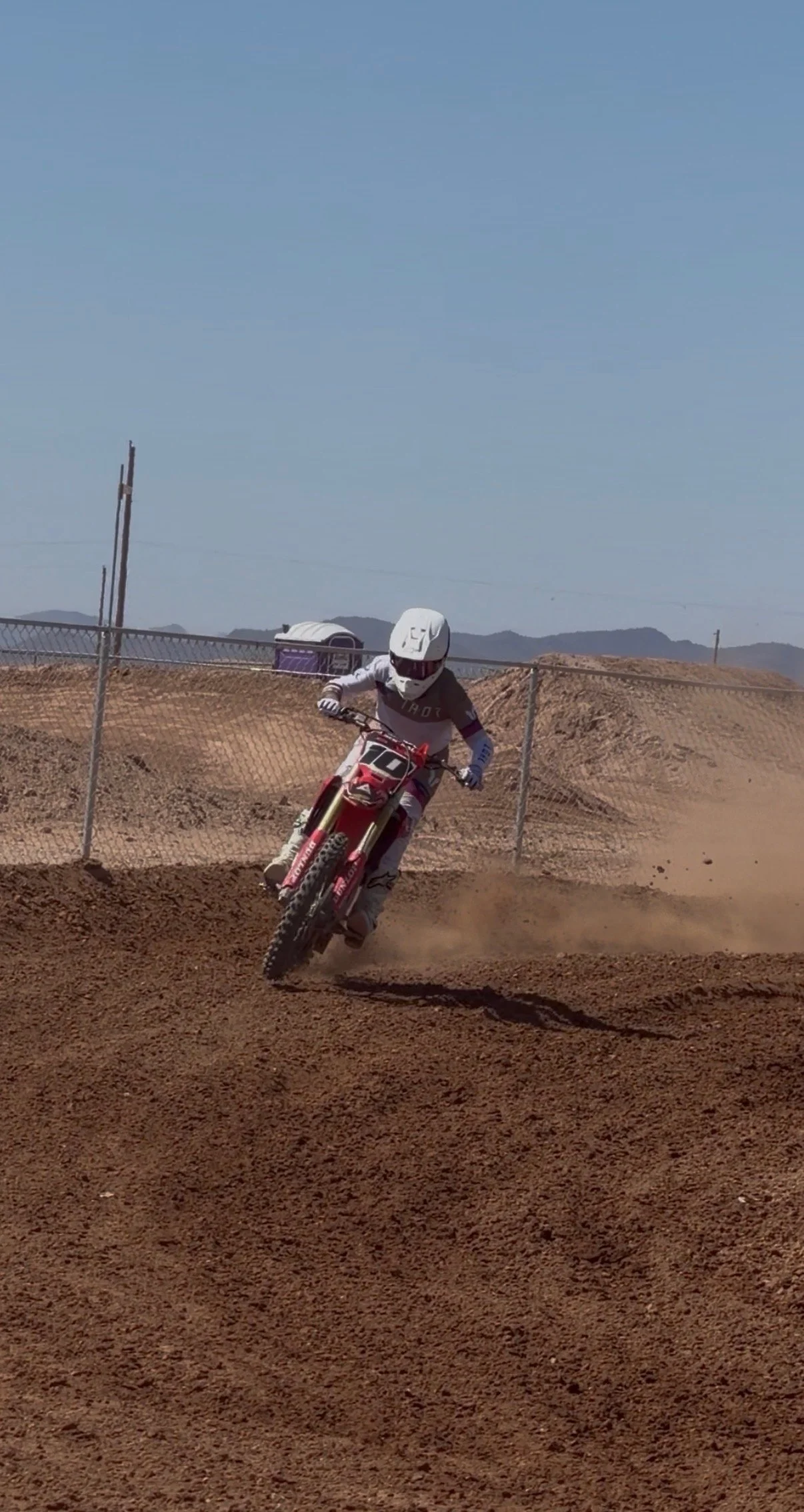 A person wearing a white helmet and riding a red dirt bike on a dirt track under a clear blue sky.