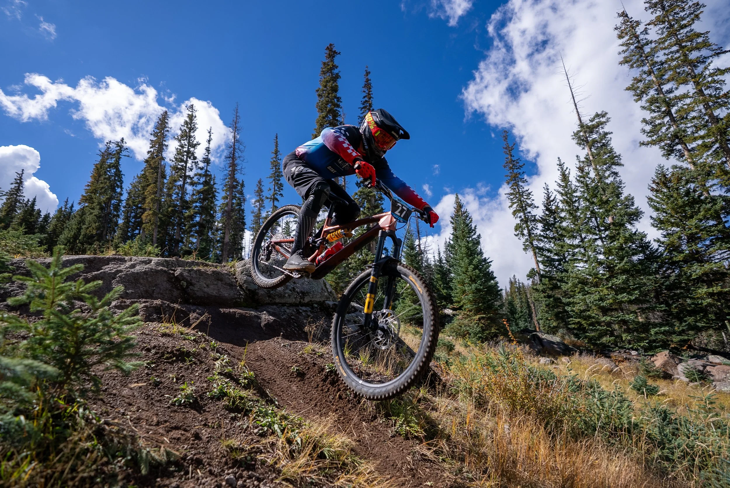 A mountain biker jumping off a rocky ledge downhill in a forested area during daytime, with tall pine trees and a blue sky with some clouds in the background.