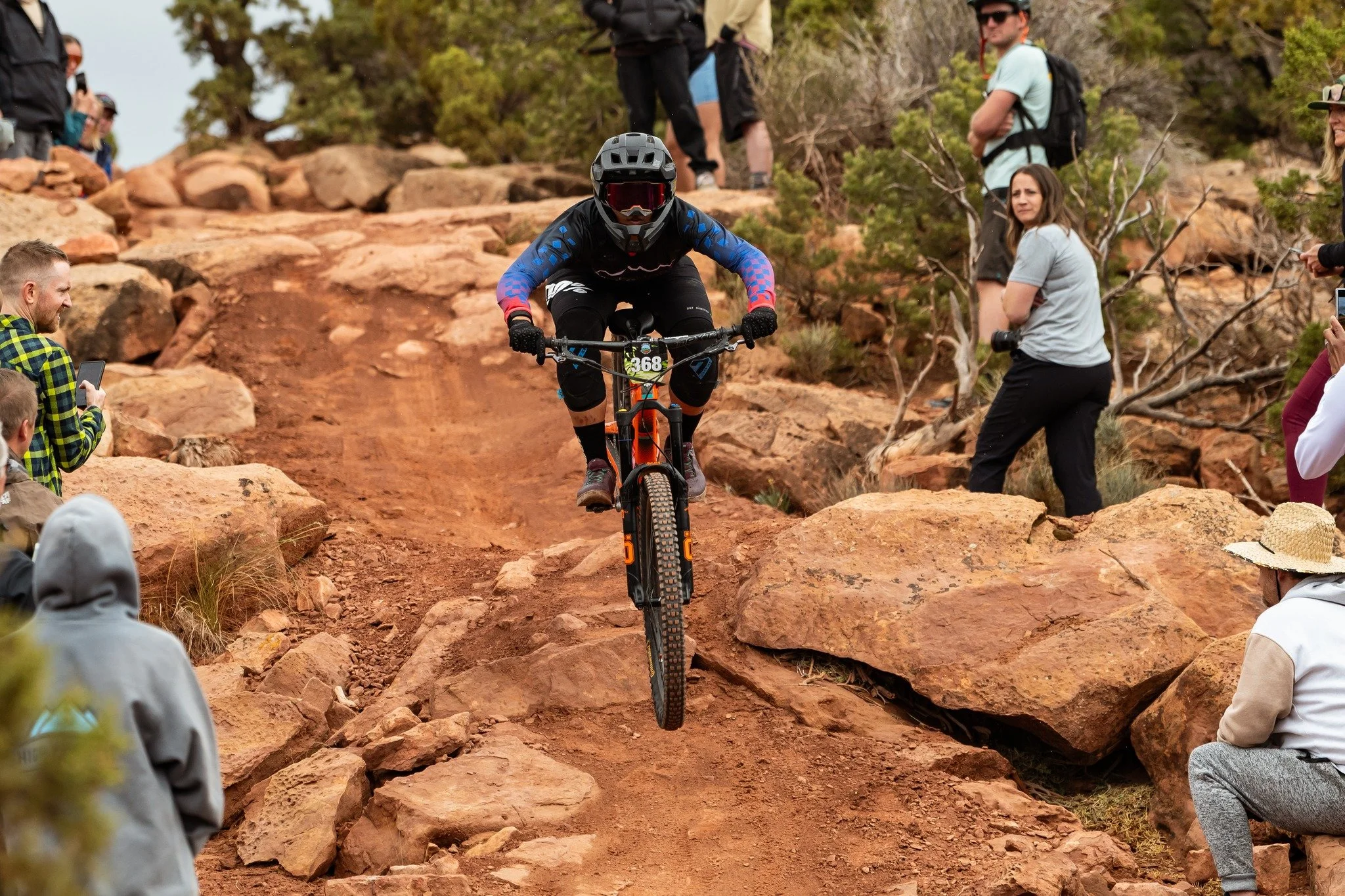 A mountain biker in black gear with a bib number 368, riding downhill on a rocky, dirt trail surrounded by spectators.