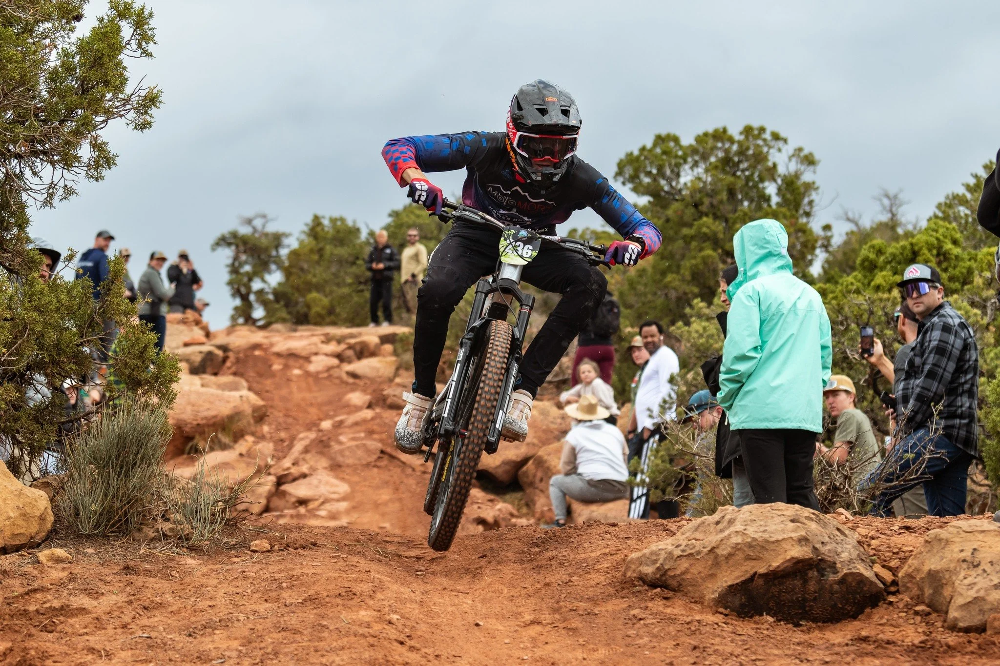 A mountain biker wearing a helmet and racing gear jumps off a dirt trail during a race as onlookers watch from the sidelines on a rocky, wooded outdoor trail.