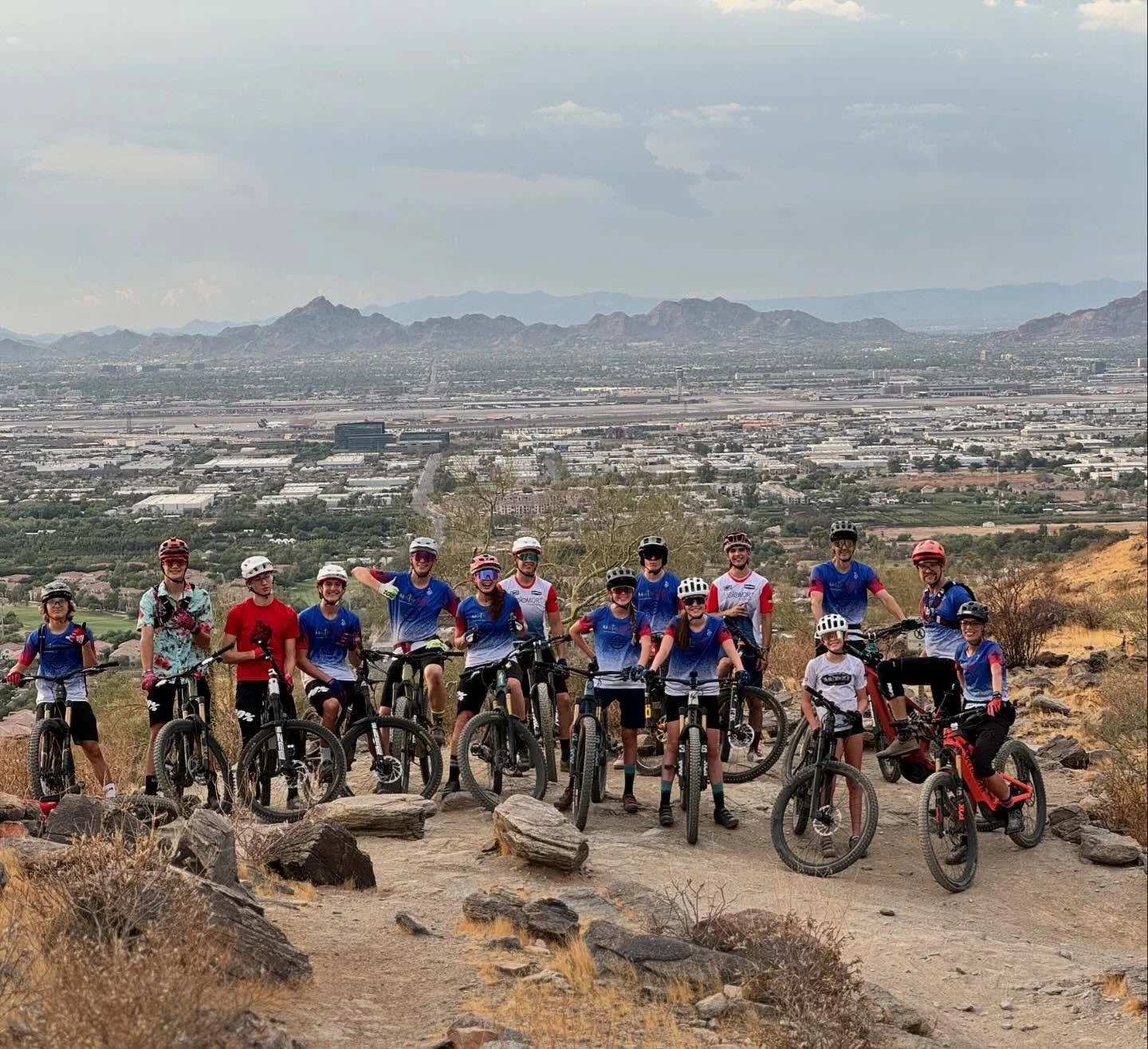 A group of children and adults on mountain bikes posed on a dirt trail with a cityscape and mountains in the background.