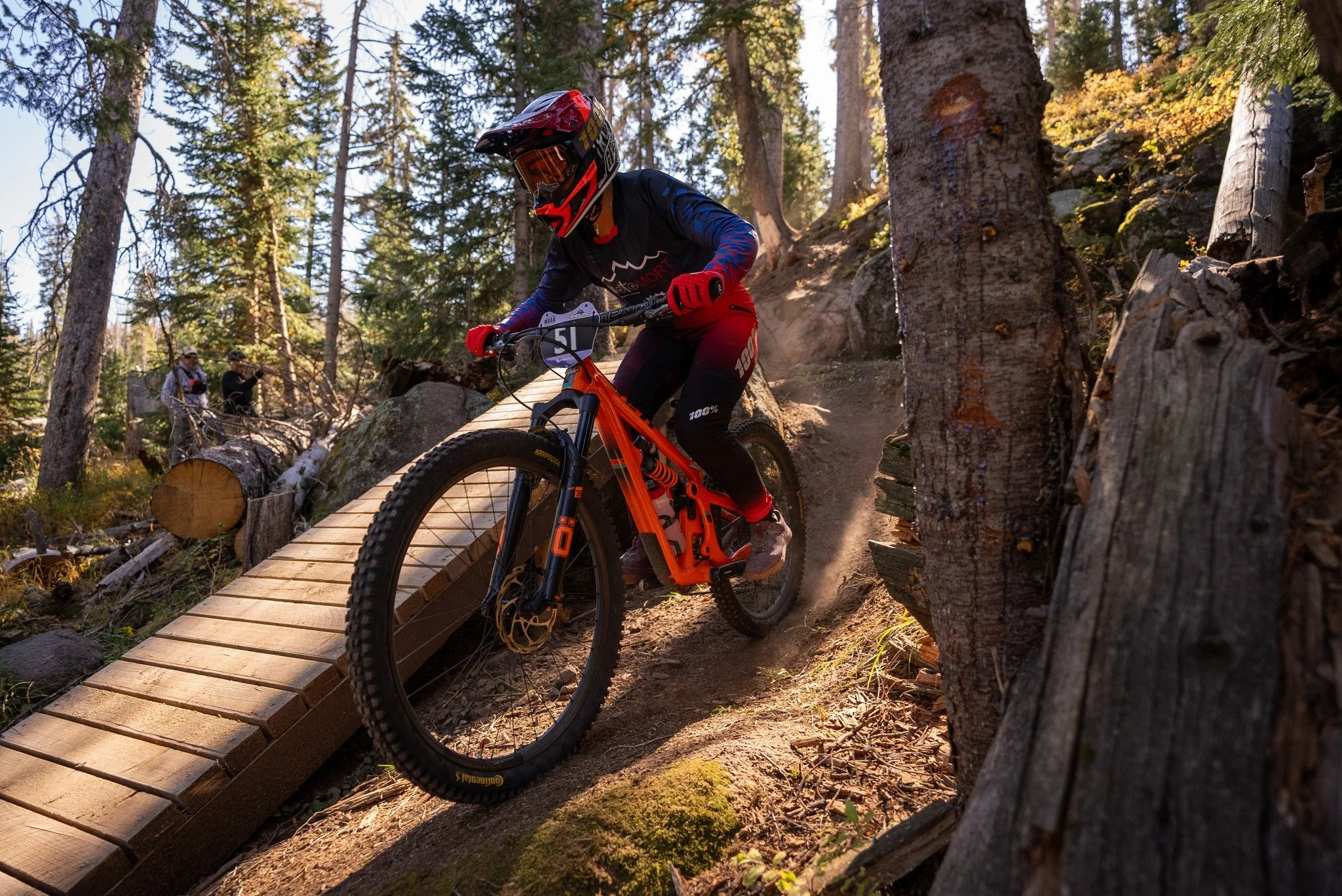 A mountain biker jumping off a wooden ramp on a dirt trail in a forest, with trees and other people in the background.