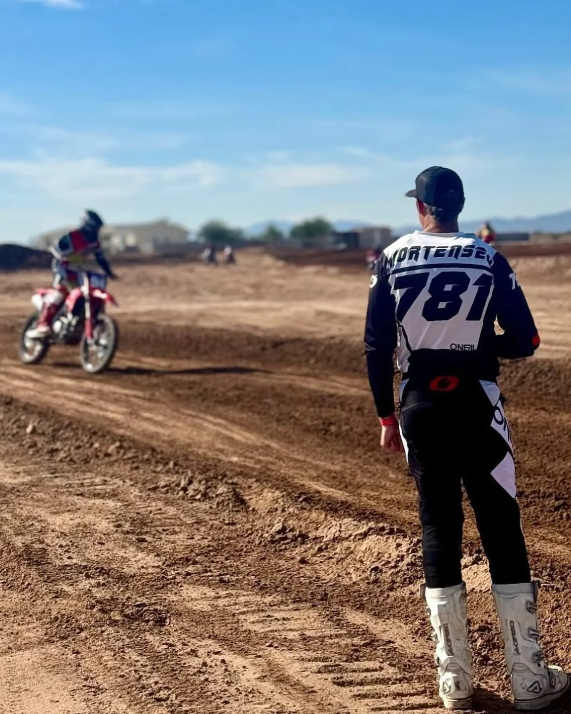 A person in motocross gear watching a dirt bike rider on a dirt track under a blue sky.