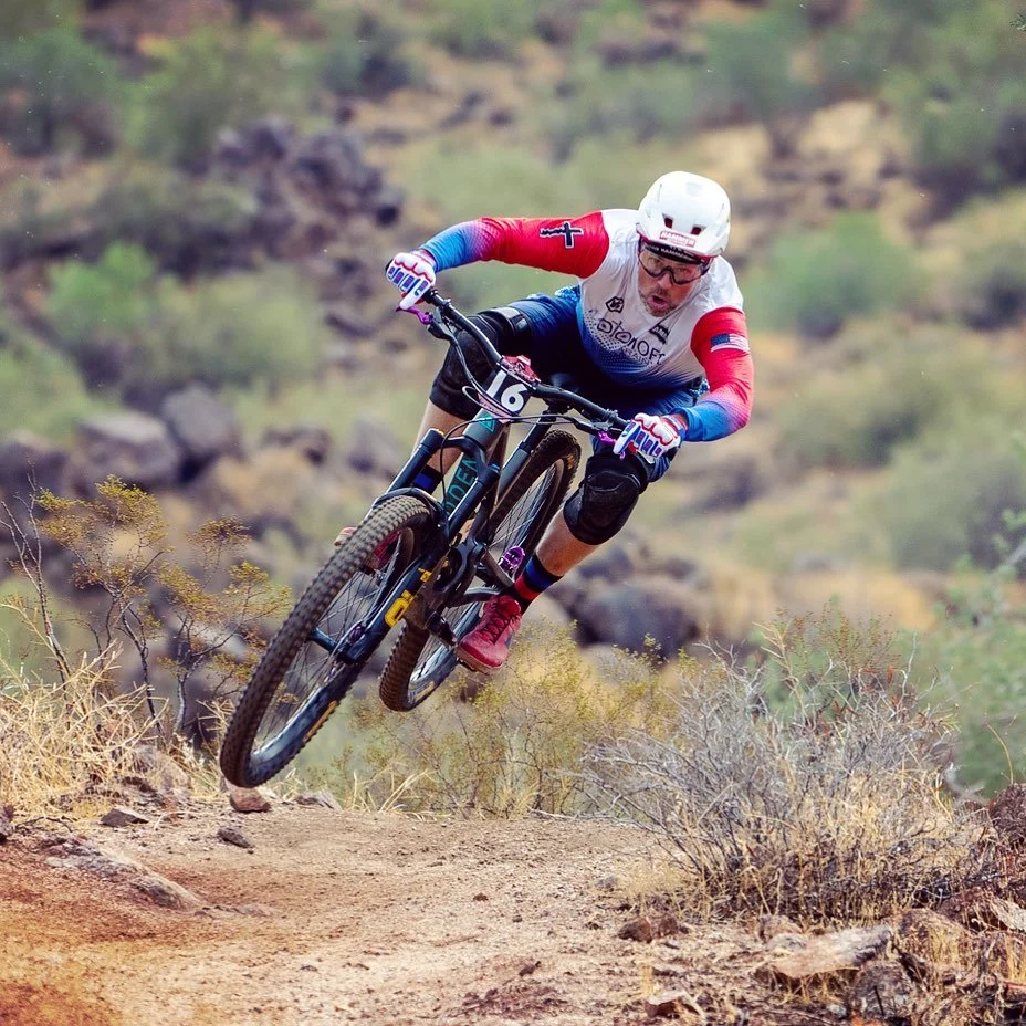 A mountain biker wearing a white helmet, glasses, a red, white, and blue jersey, black shorts, and red shoes, is mid-air while riding a black mountain bike on a dirt trail surrounded by desert bushes and rocks.