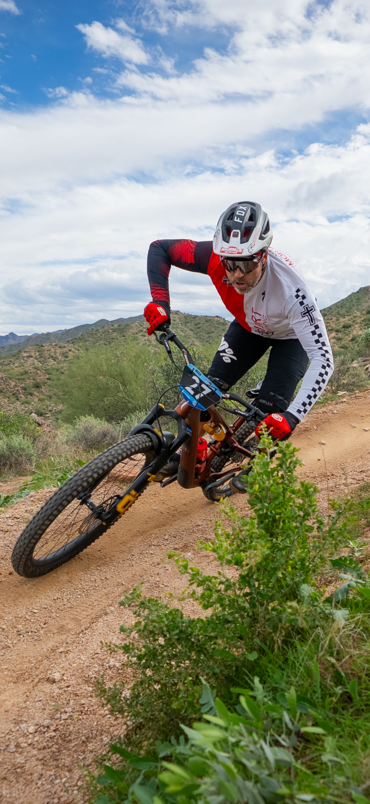 A mountain biker wearing a helmet, sunglasses, red gloves, and a white and red jersey riding on a dirt trail through a desert landscape with mountains and a partly cloudy sky in the background.