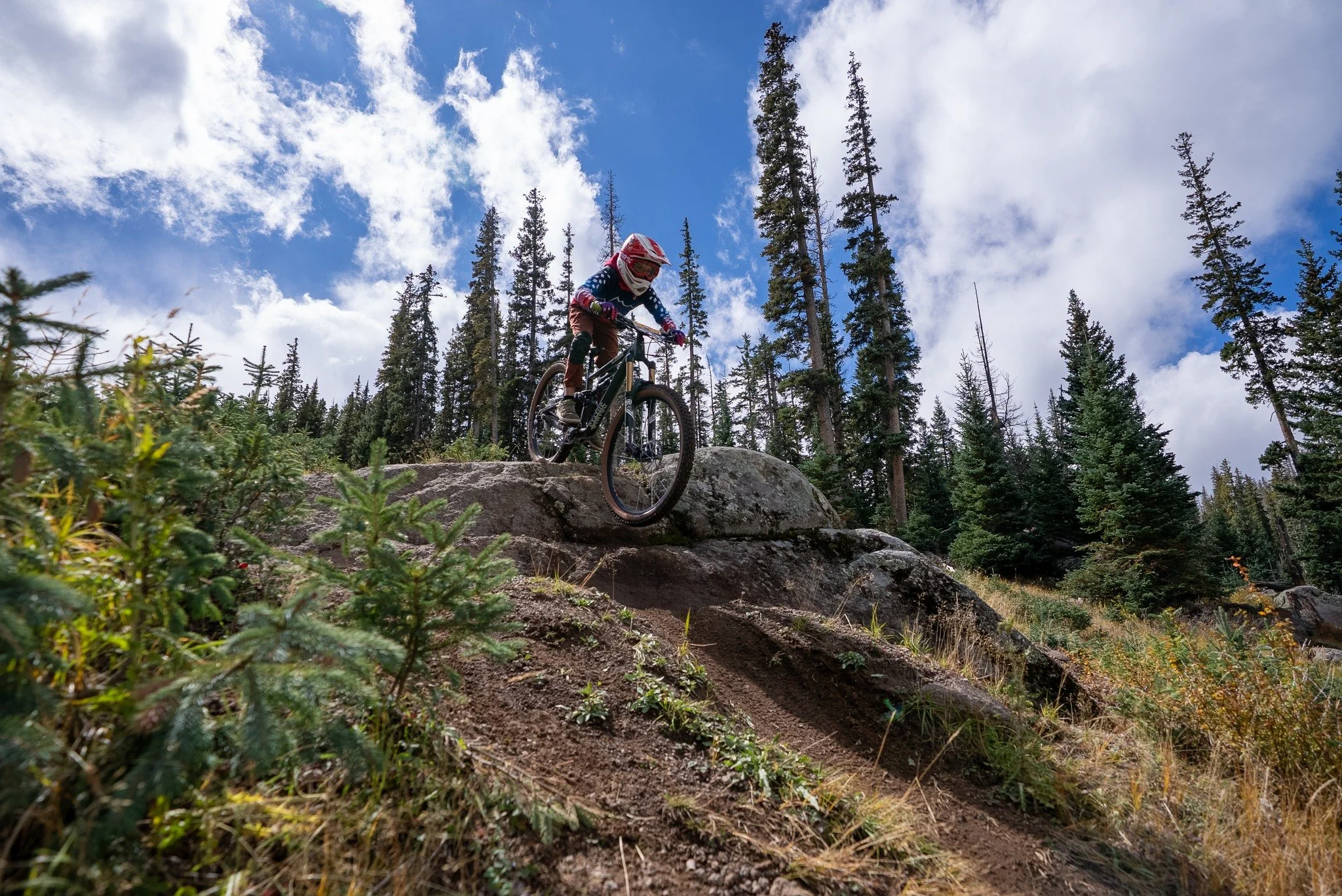 A person mountain biking downhill on a rocky trail in a forested area under a partly cloudy sky.