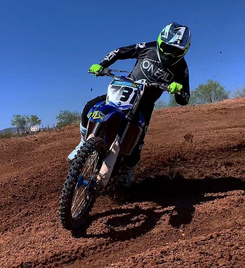 A motocross rider wearing a black suit with green gloves and a yellow and black helmet rides a blue dirt bike on a dirt trail under a clear blue sky.