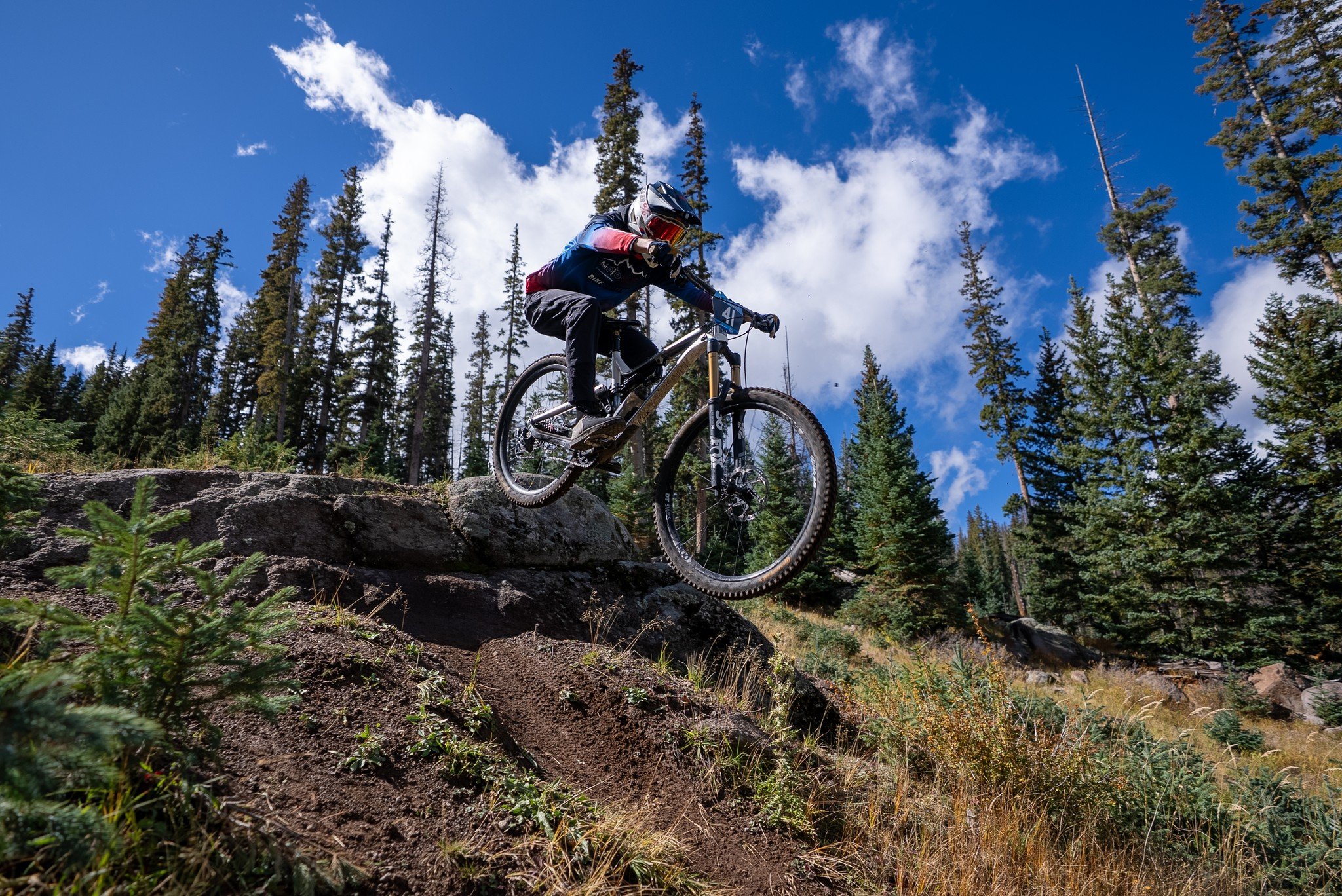 Mountain biker wearing helmet and protective gear riding down a rocky hillside surrounded by pine trees under a blue sky with clouds.