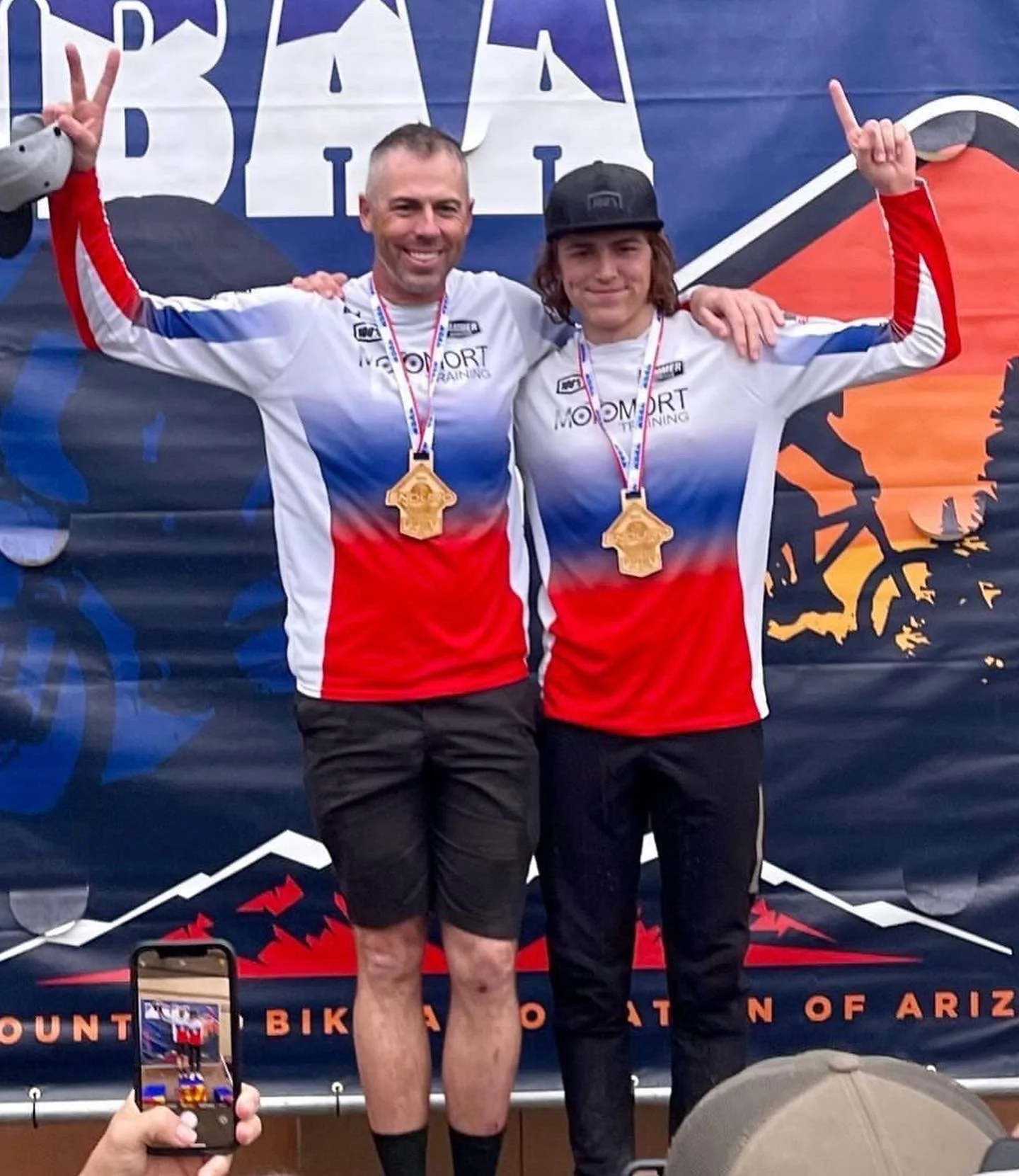 Two men standing side by side on a podium, wearing matching blue, red, and white cycling jerseys, each with a gold medal around their neck, smiling and making victory signs with their hands. They are celebrating a biking event at the Mount Lemmon Bic