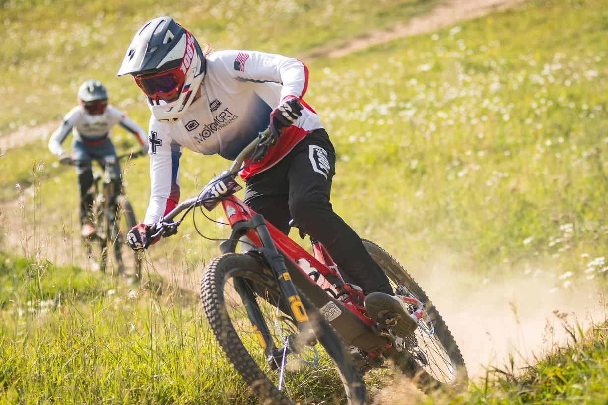 Two mountain bikers riding on a grassy trail with the lead biker in a white jersey and helmet, kicking up dust.
