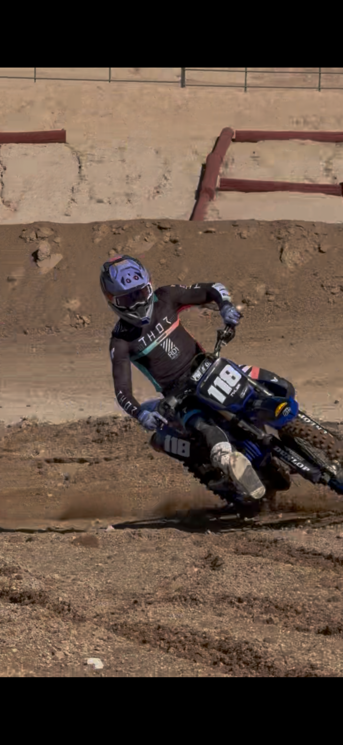 A motocross rider wearing a black helmet, goggles, and gear, riding a dirt bike on a rugged dirt track.